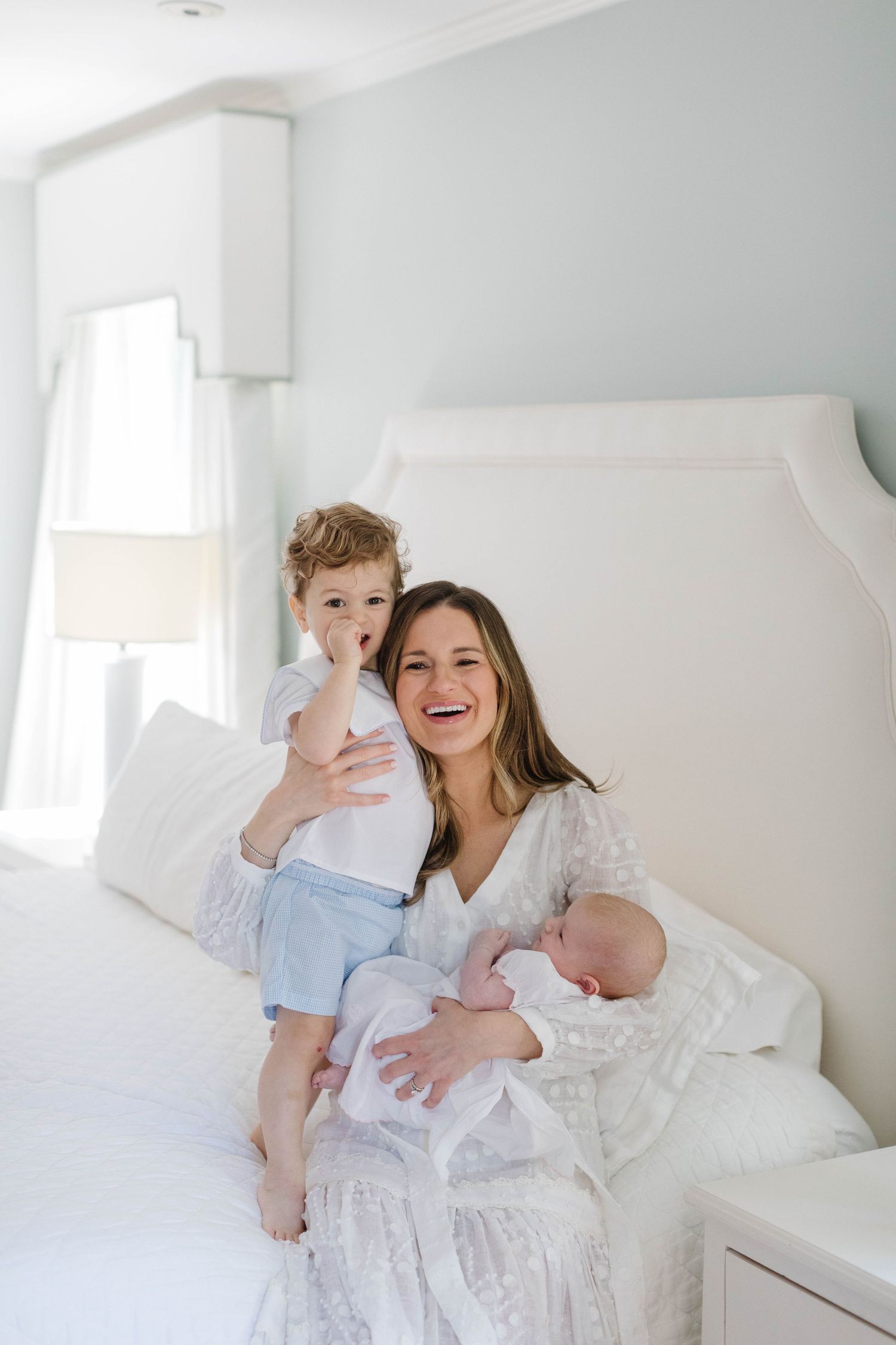 A joyful family moment in a bright white bedroom with a toddler and newborn.