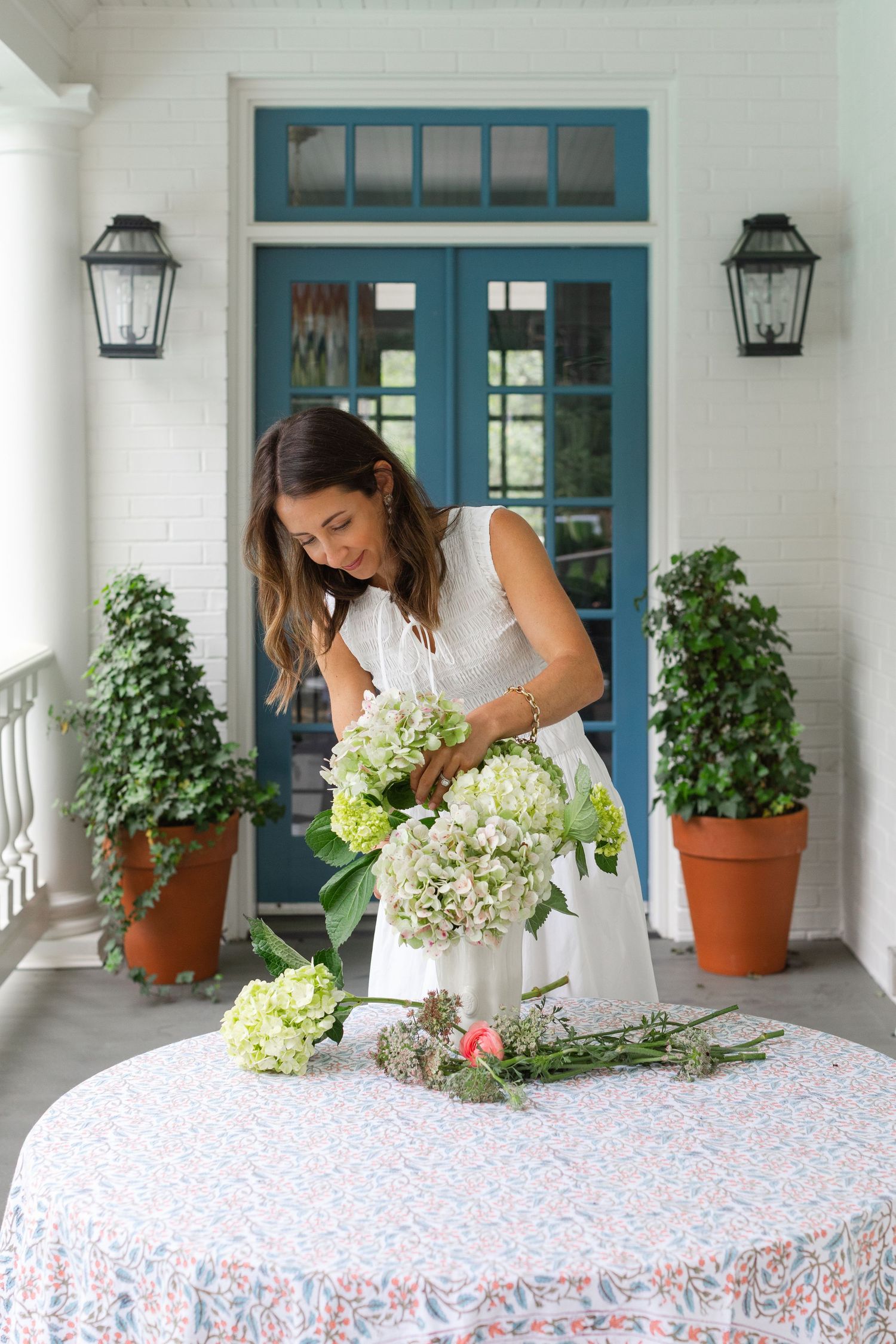 Woman arranging white hydrangeas in a vase on a porch table with a floral tablecloth and blue double doors.