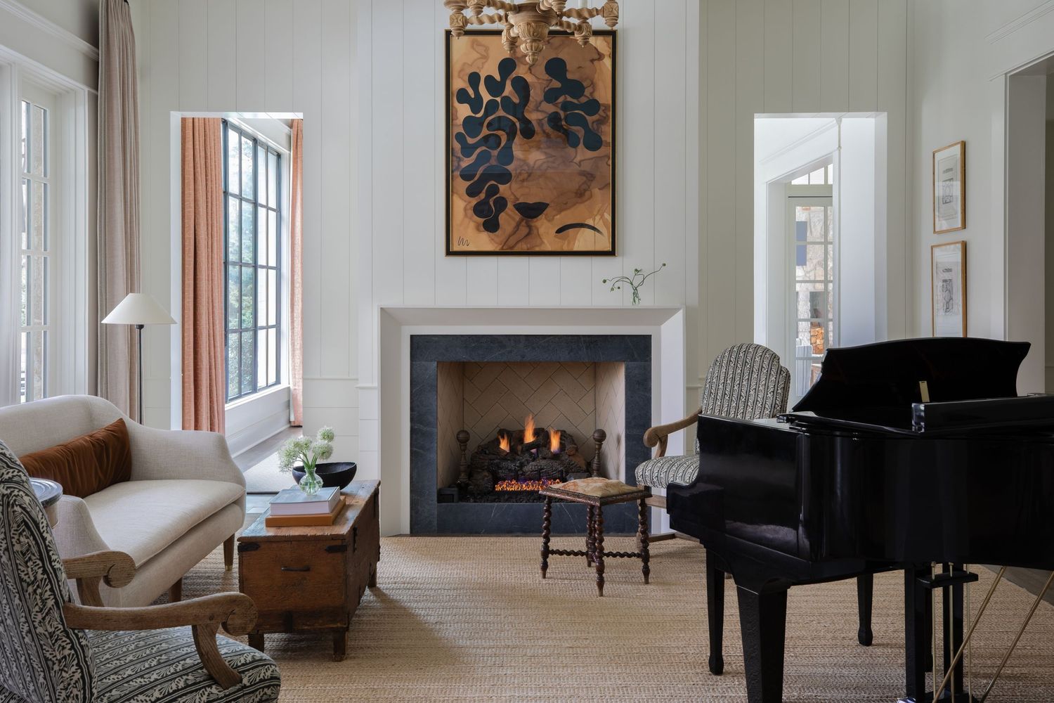 Elegant living room with a grand piano, lit fireplace, botanical artwork, and classic armchairs on a neutral carpet.