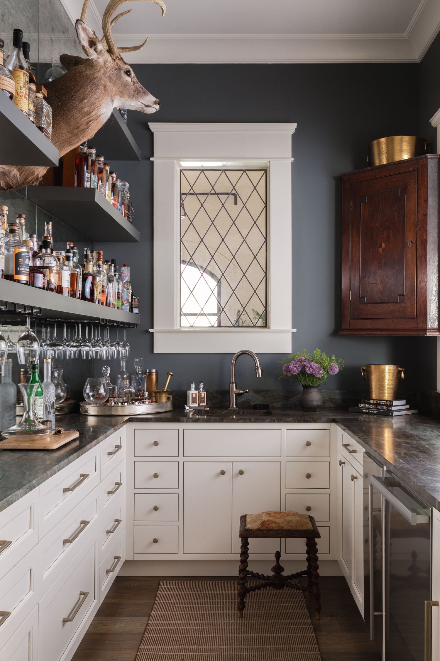 Dark moody butler's pantry with white cabinets, dark countertops, open shelving with bottles, and a leaded glass window.