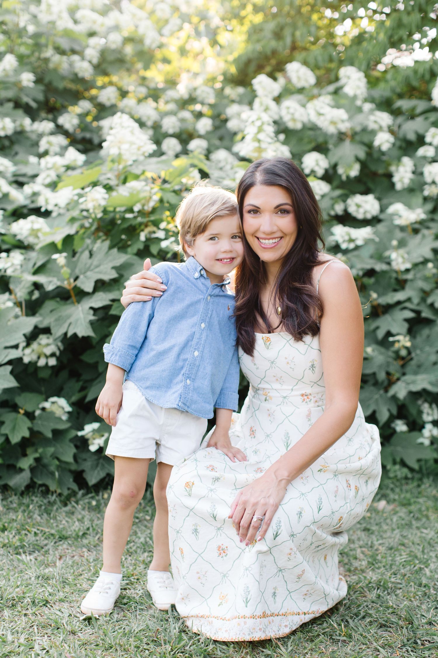 A smiling woman in a floral dress crouches beside a young boy in a blue shirt and white shorts outdoors.