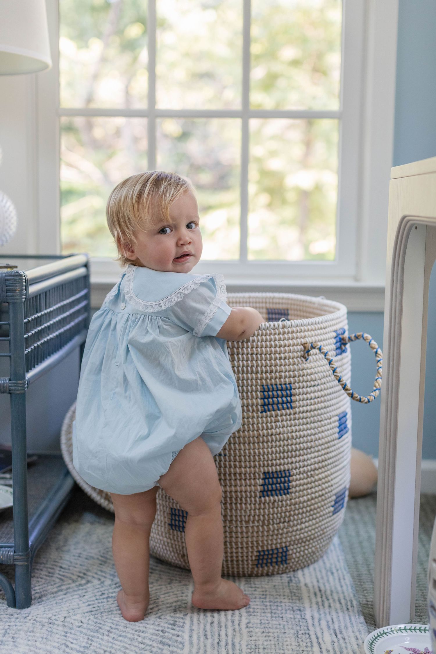 Small child in light blue dress plays near a woven storage basket by a sunny window.