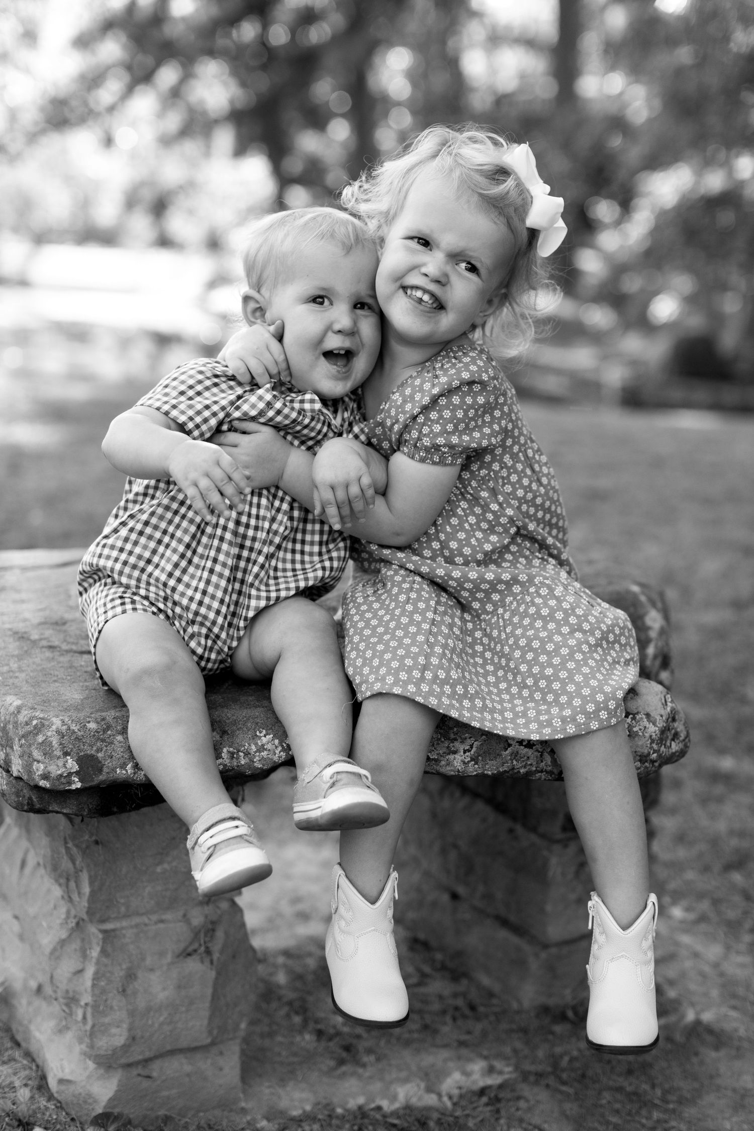 Two children sit together on a rock in a park, wearing polka dot and gingham dresses in this vintage black and white photo.