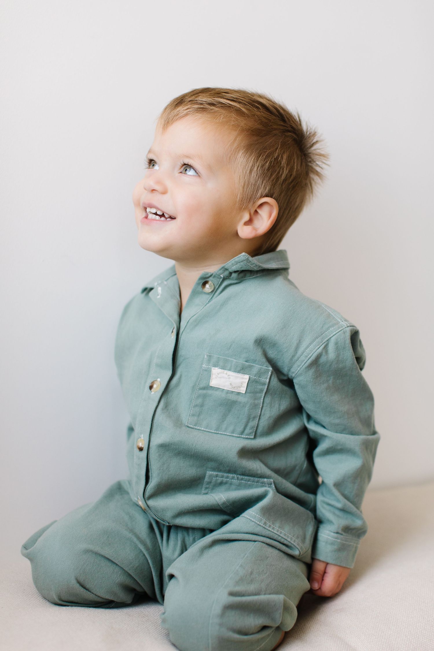 A small child wearing a sage green jumpsuit sits and smiles against a white backdrop in three candid poses.