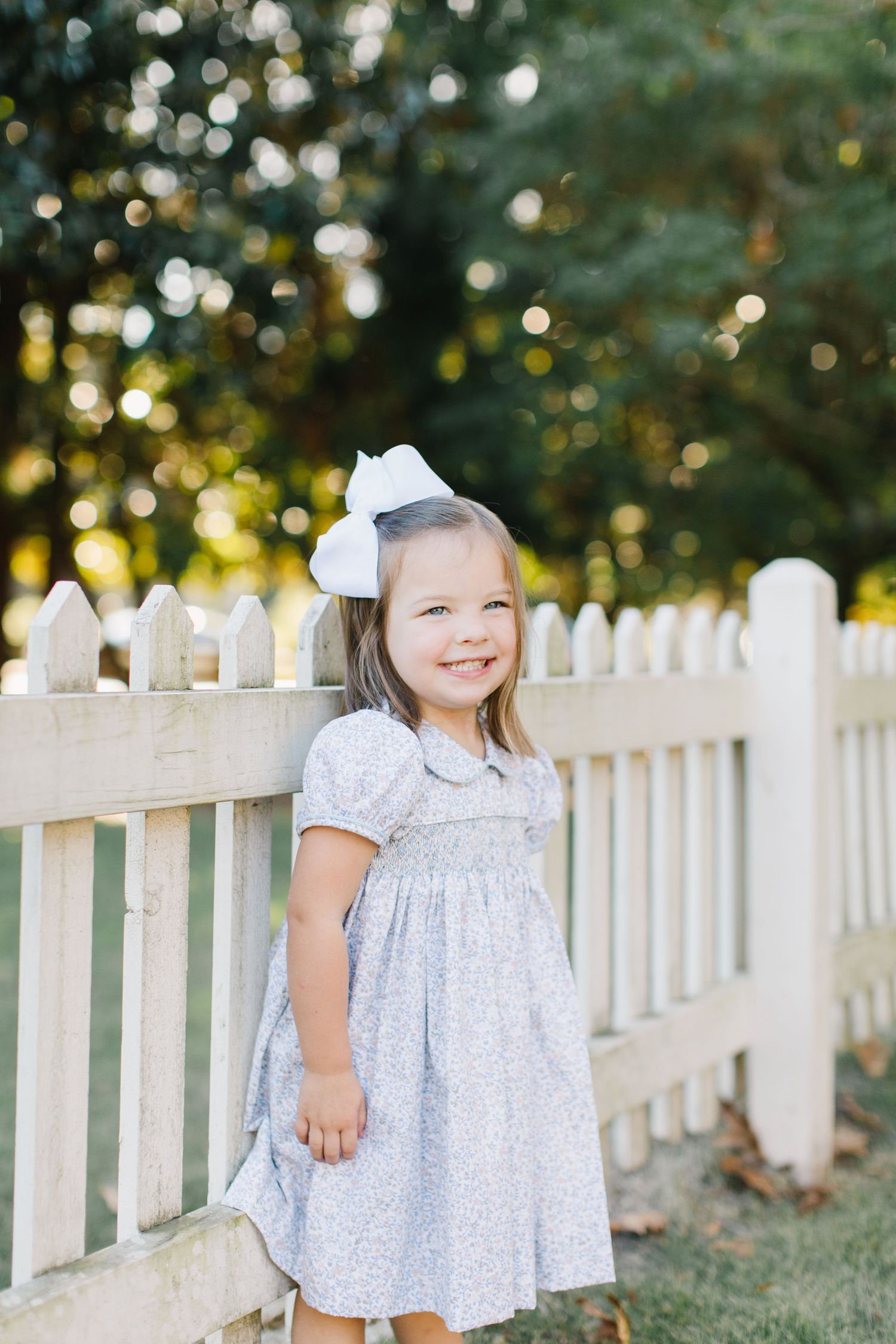 Children in matching white floral dresses pose by a white picket fence during a sunny outdoor photoshoot.