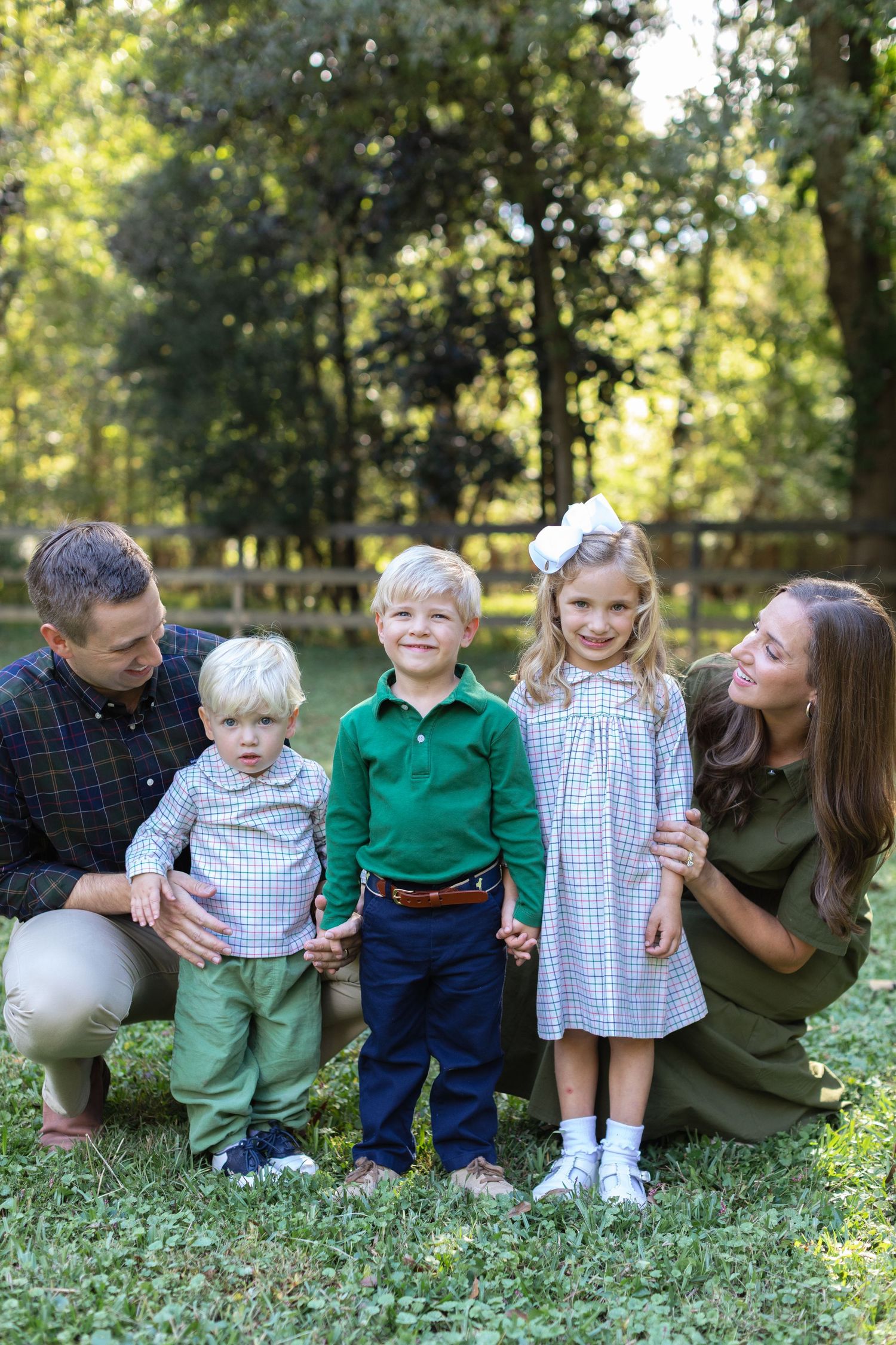 A happy family group smiles together during an outdoor photo session in a sunlit wooded setting.