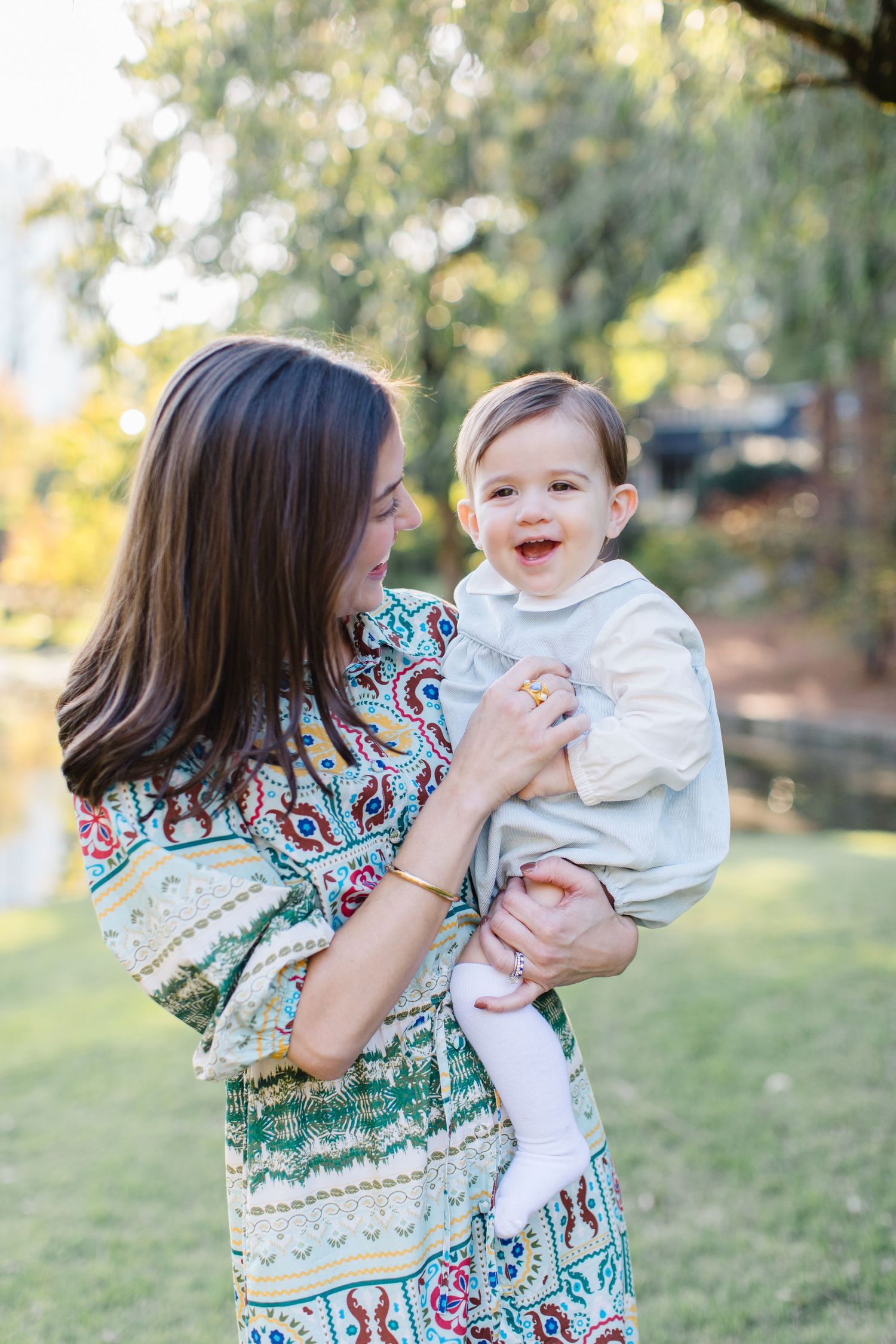 A woman in a patterned dress holds a smiling baby wearing white outdoors in a sunny garden setting.