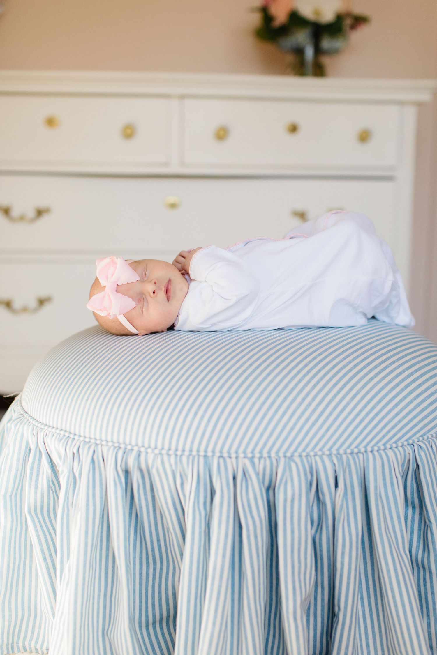 A sleeping newborn in white clothing rests on a blue and white striped ottoman near a dresser.