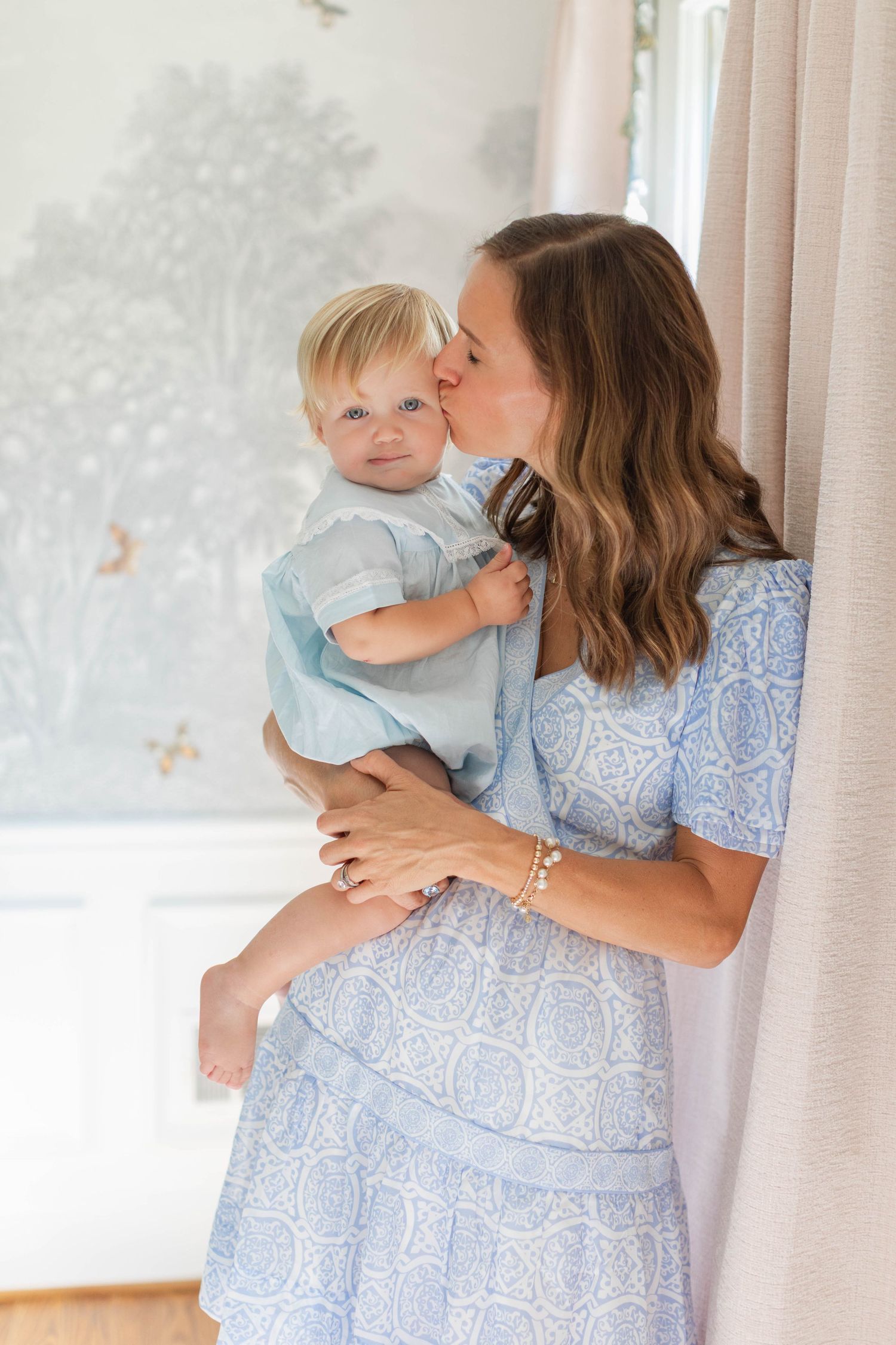 Mother and baby in matching light blue floral dresses share tender moments by window with sheer curtains.