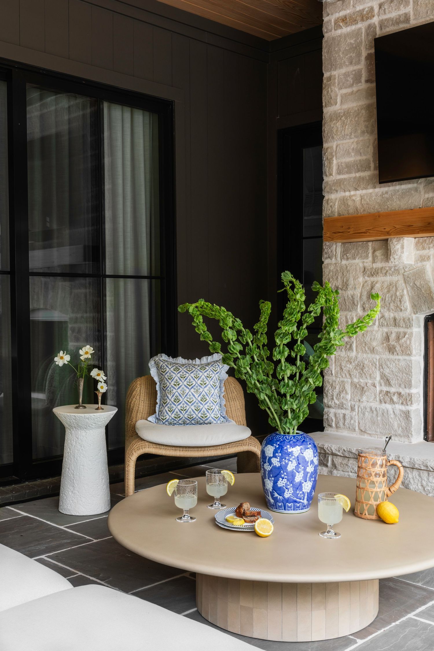 Outdoor patio with round wooden coffee table, blue ginger jar vase, wicker chair, stone fireplace, and refreshing lemonade drinks.