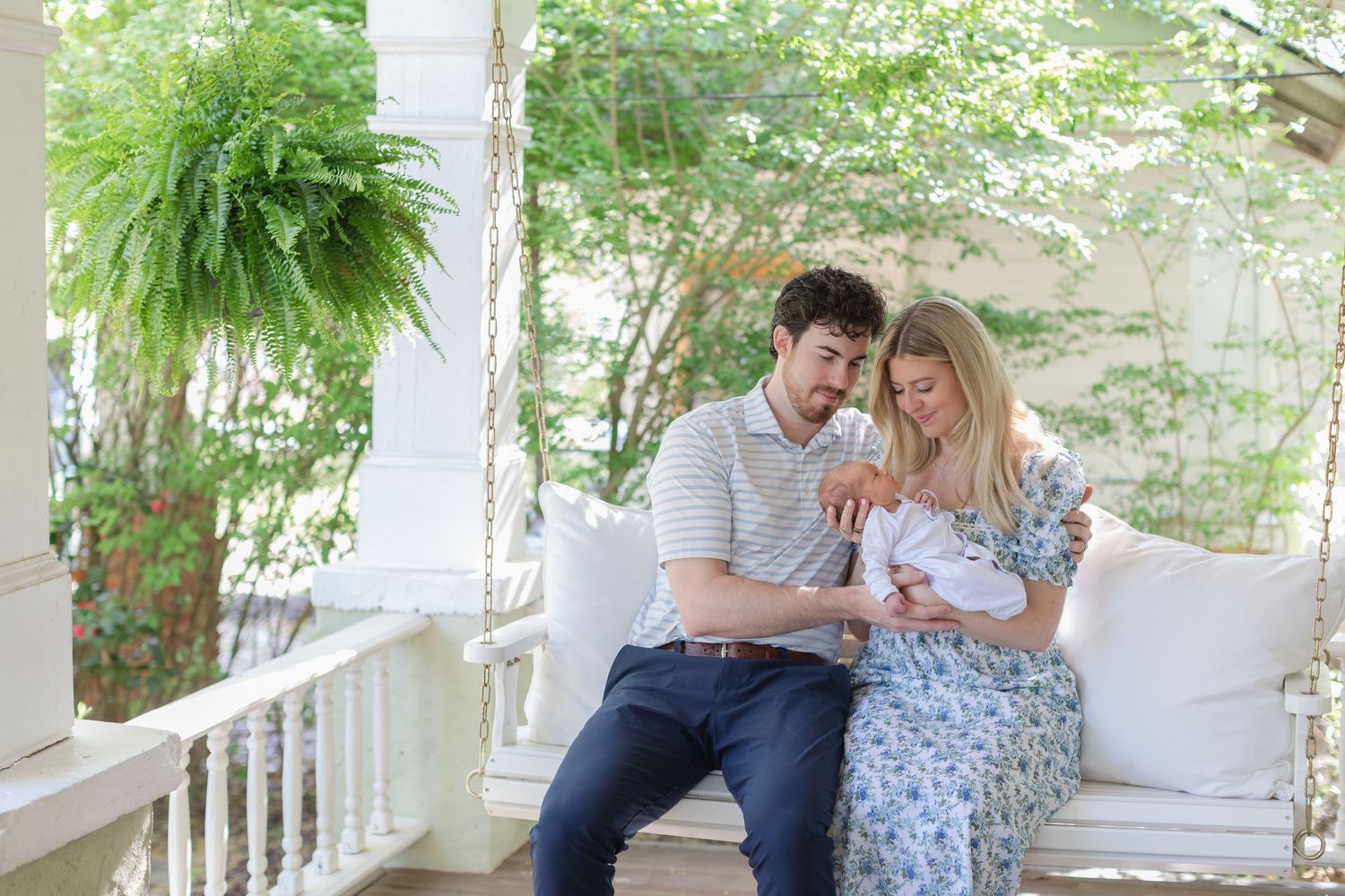 New parents sit on a porch swing admiring their newborn baby surrounded by lush green trees.