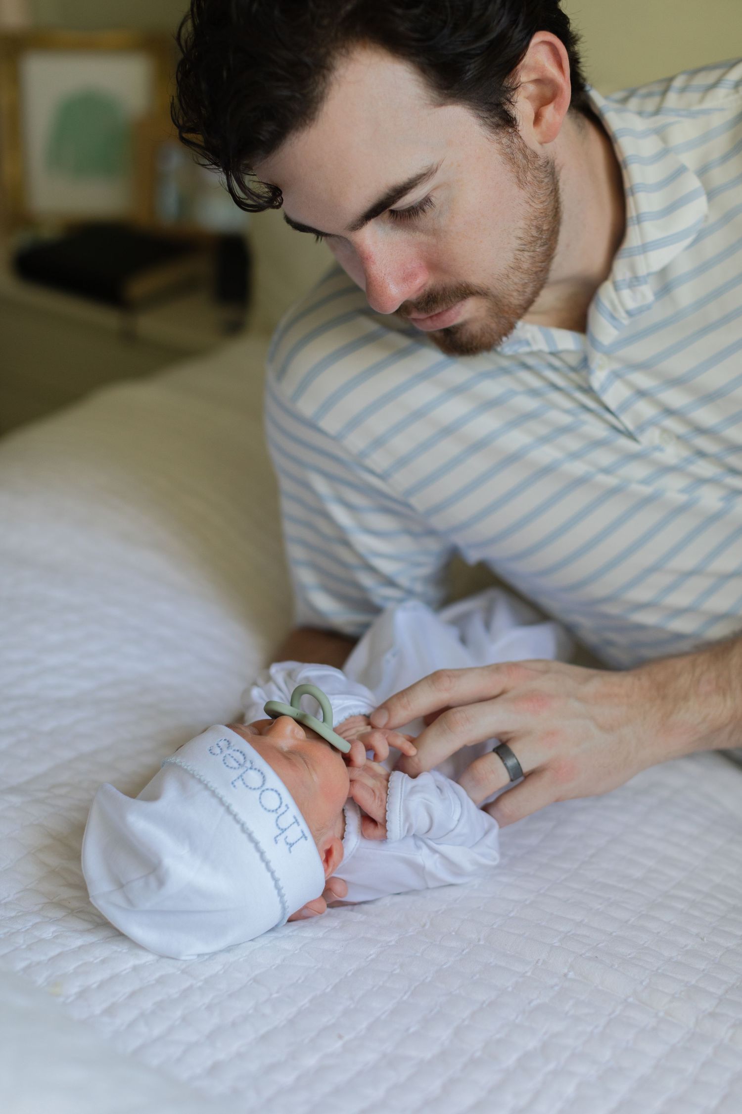 Father in striped shirt gazing lovingly at newborn baby wearing white hat on white quilted bed.