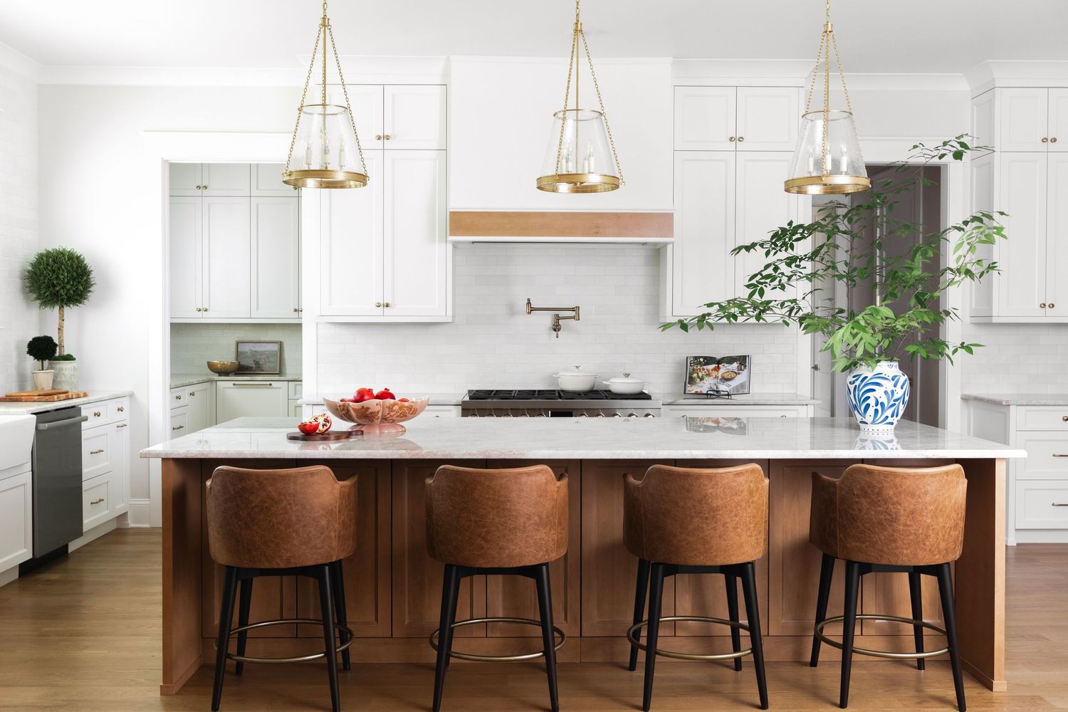 Modern white kitchen with gold pendant lights, wood island, leather bar stools, marble countertops, and fresh greenery accents.
