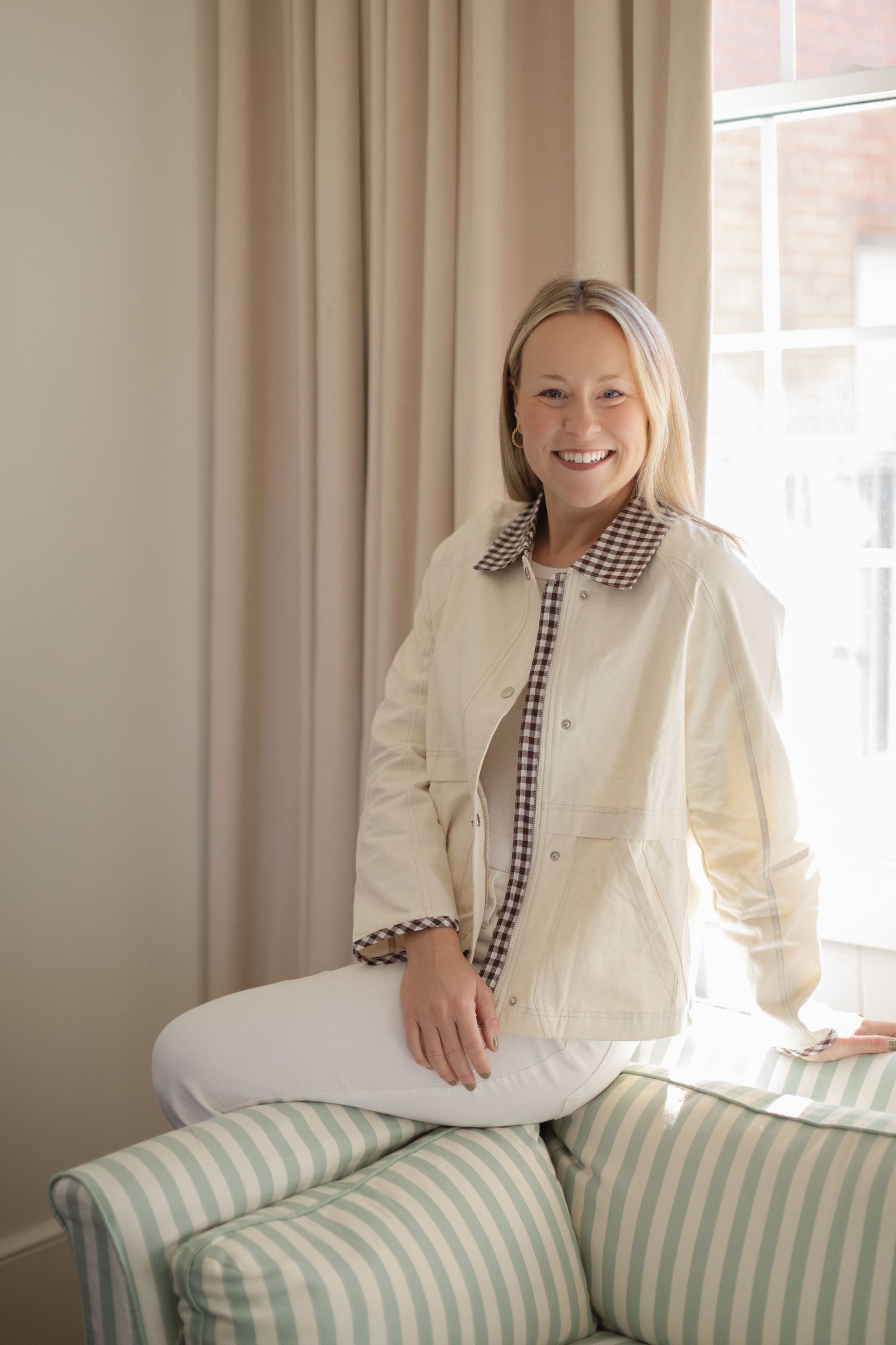 Smiling blonde woman in cream jacket and white jeans sitting on striped chair near window.