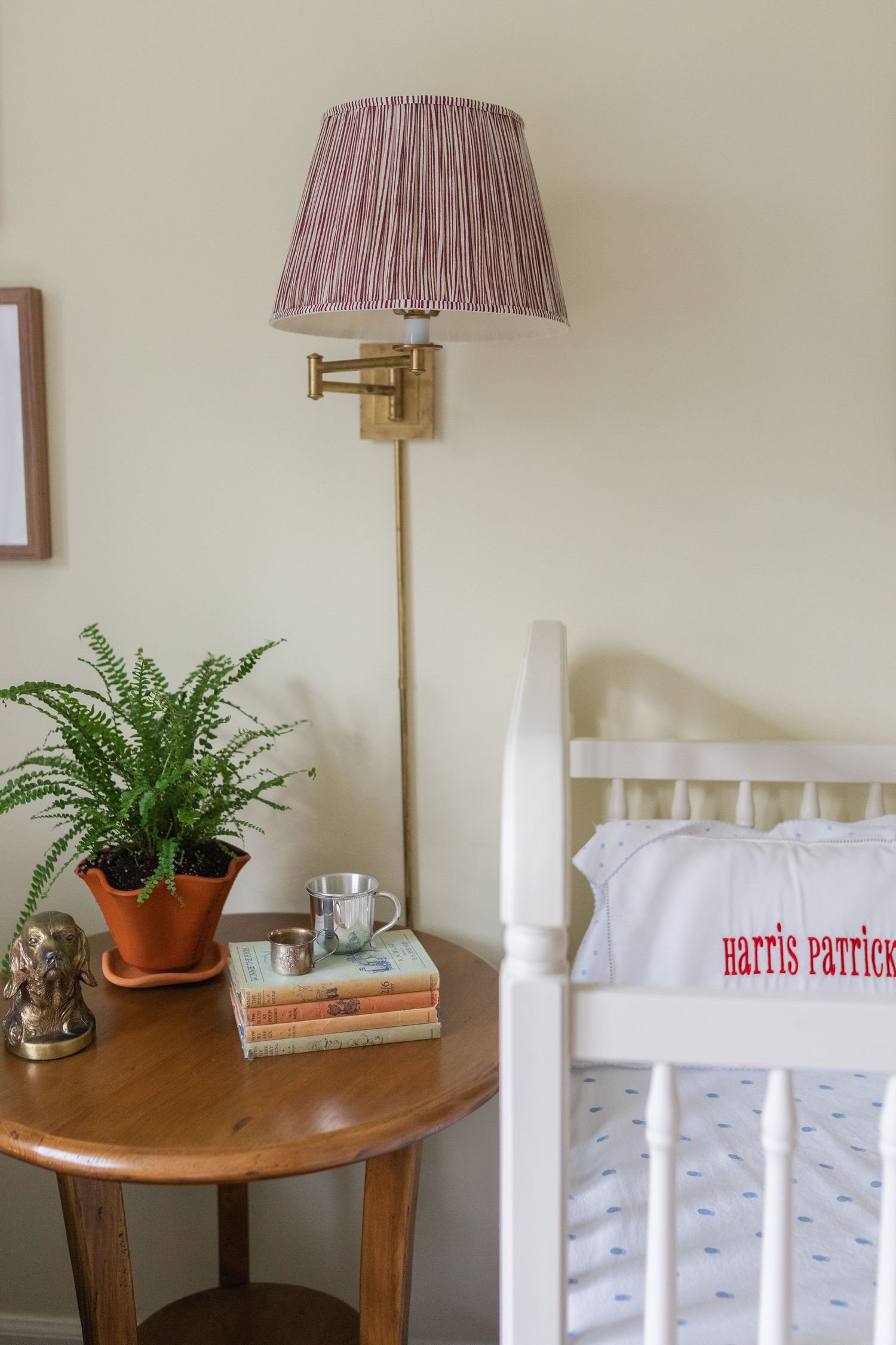 Cozy bedroom corner with brass wall lamp, fern plant, wooden side table and white crib with embroidered pillow.