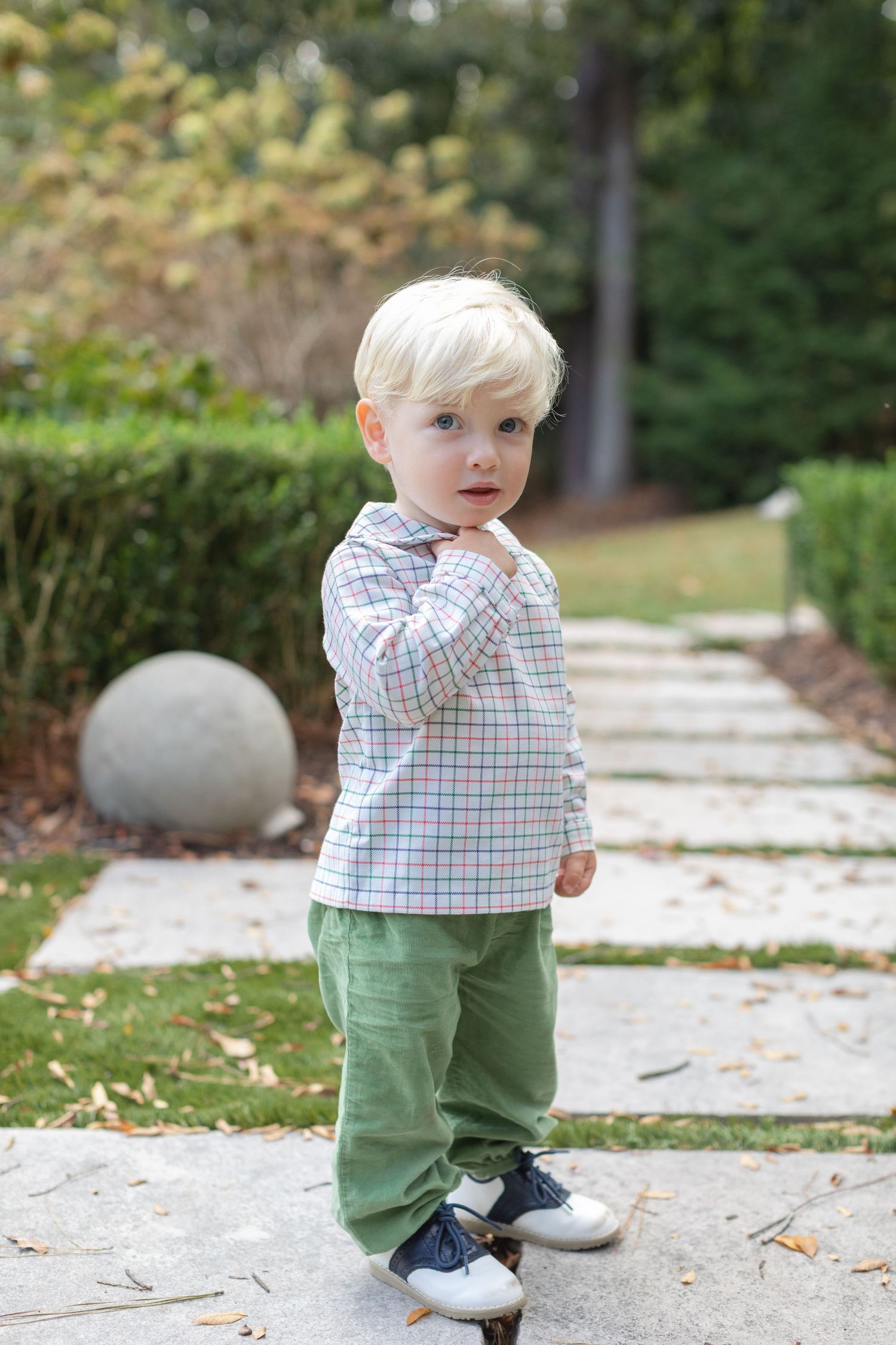 A toddler in a plaid shirt and green pants stands on a stone pathway in a garden setting.