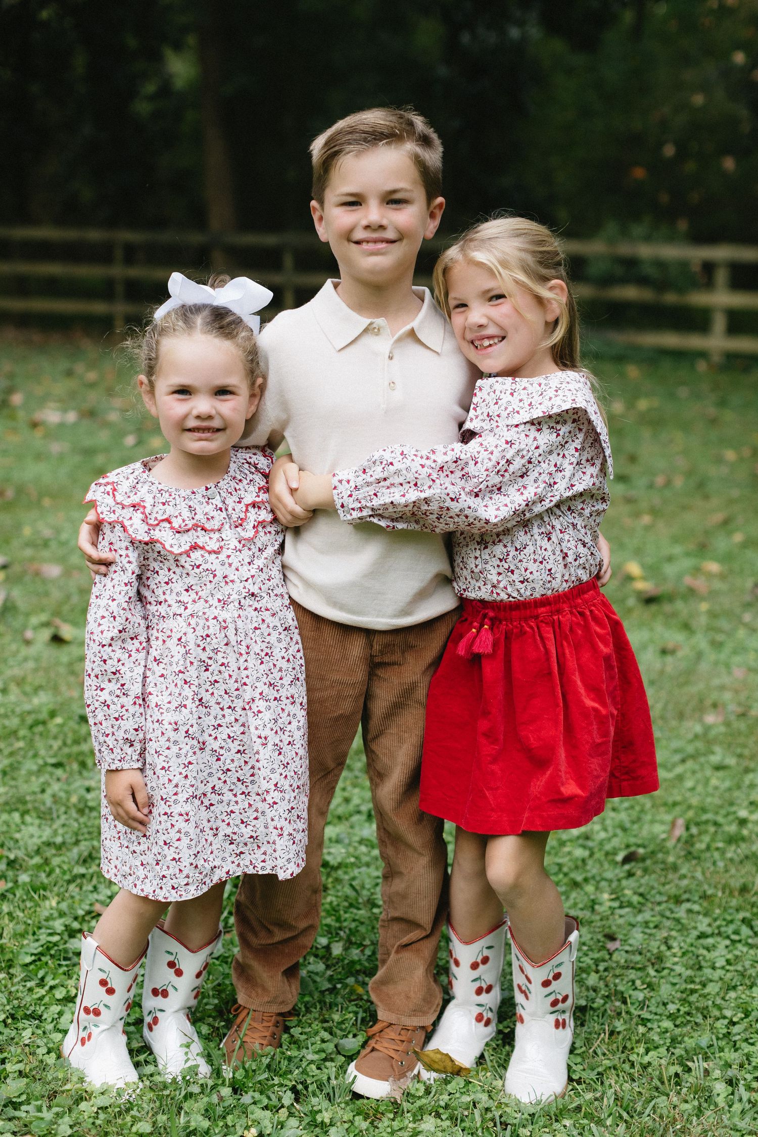 Three siblings in coordinating outfits of red, brown and floral prints pose together on green grass.