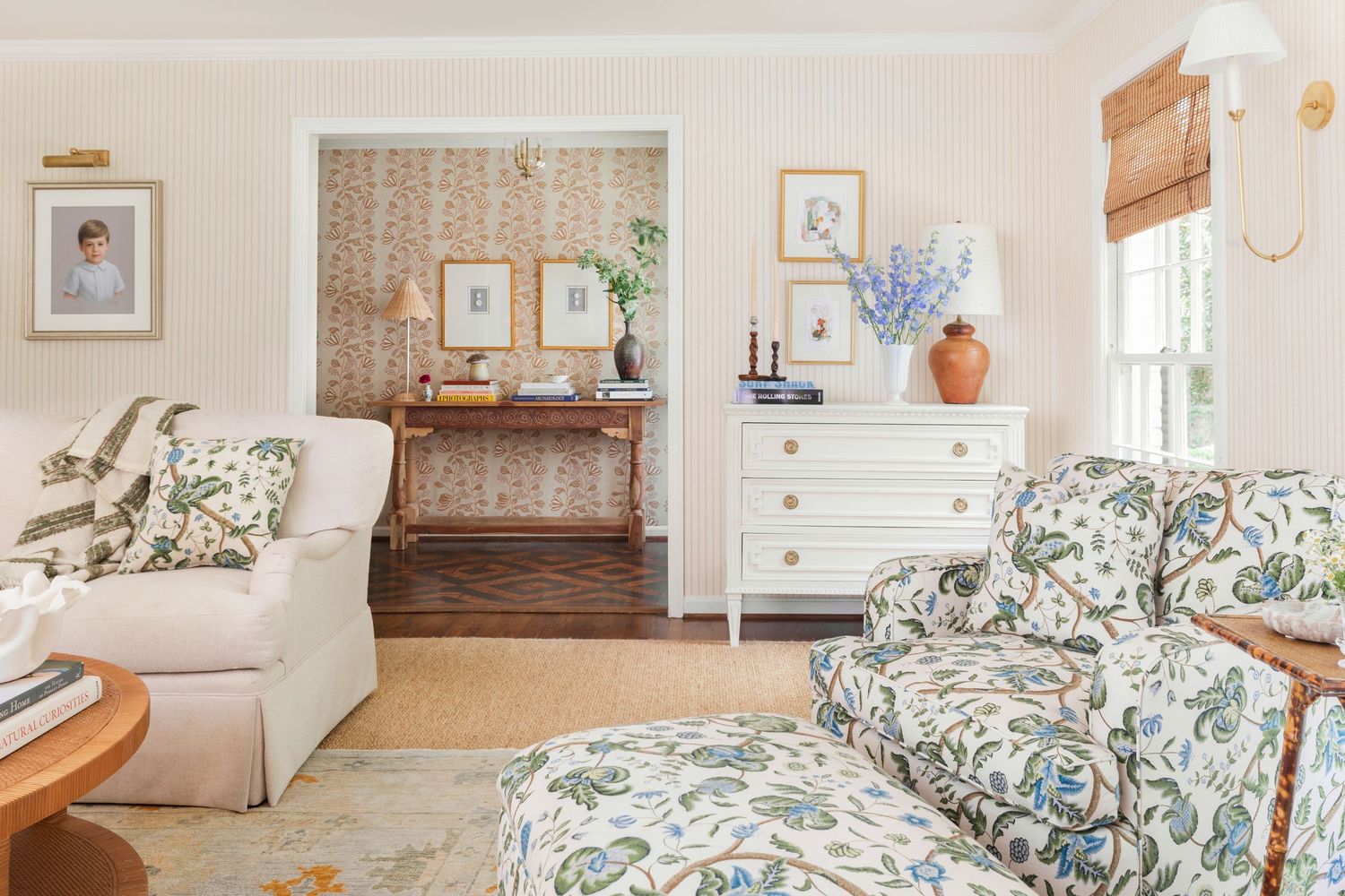 Cozy living room with floral armchair, white dresser, patterned wallpaper, and blue hydrangeas in a warm, cottage-style decor.