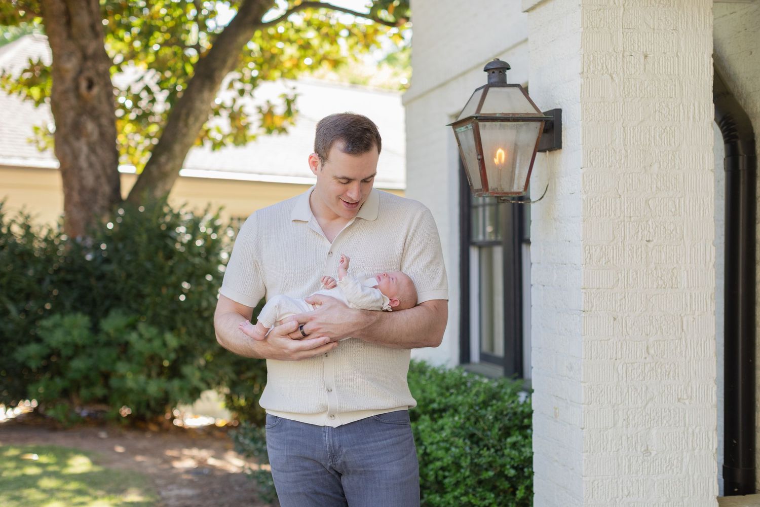 A father tenderly holds his newborn baby outside a elegant white home surrounded by lush green trees.