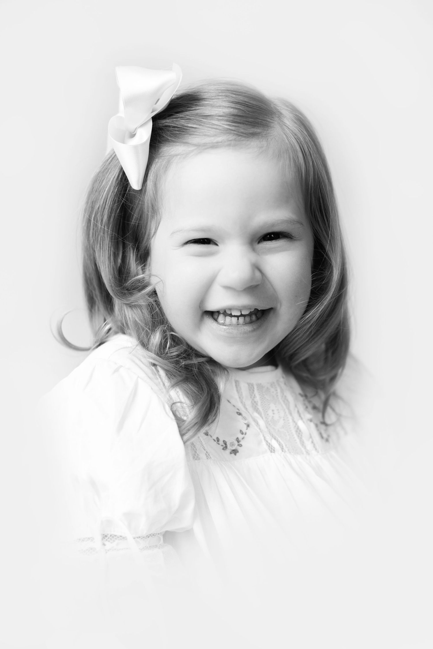 Black and white portrait of a joyful little girl with a bow in her hair, wearing a white dress, smiling brightly.