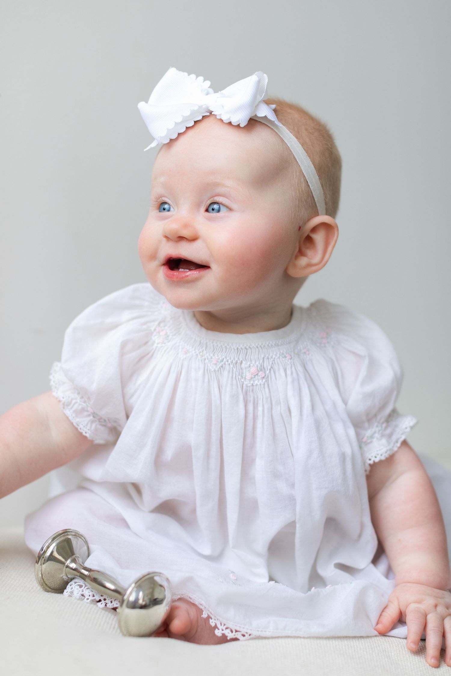Smiling baby girl in white smocked dress and bow headband sitting with silver rattle toy.
