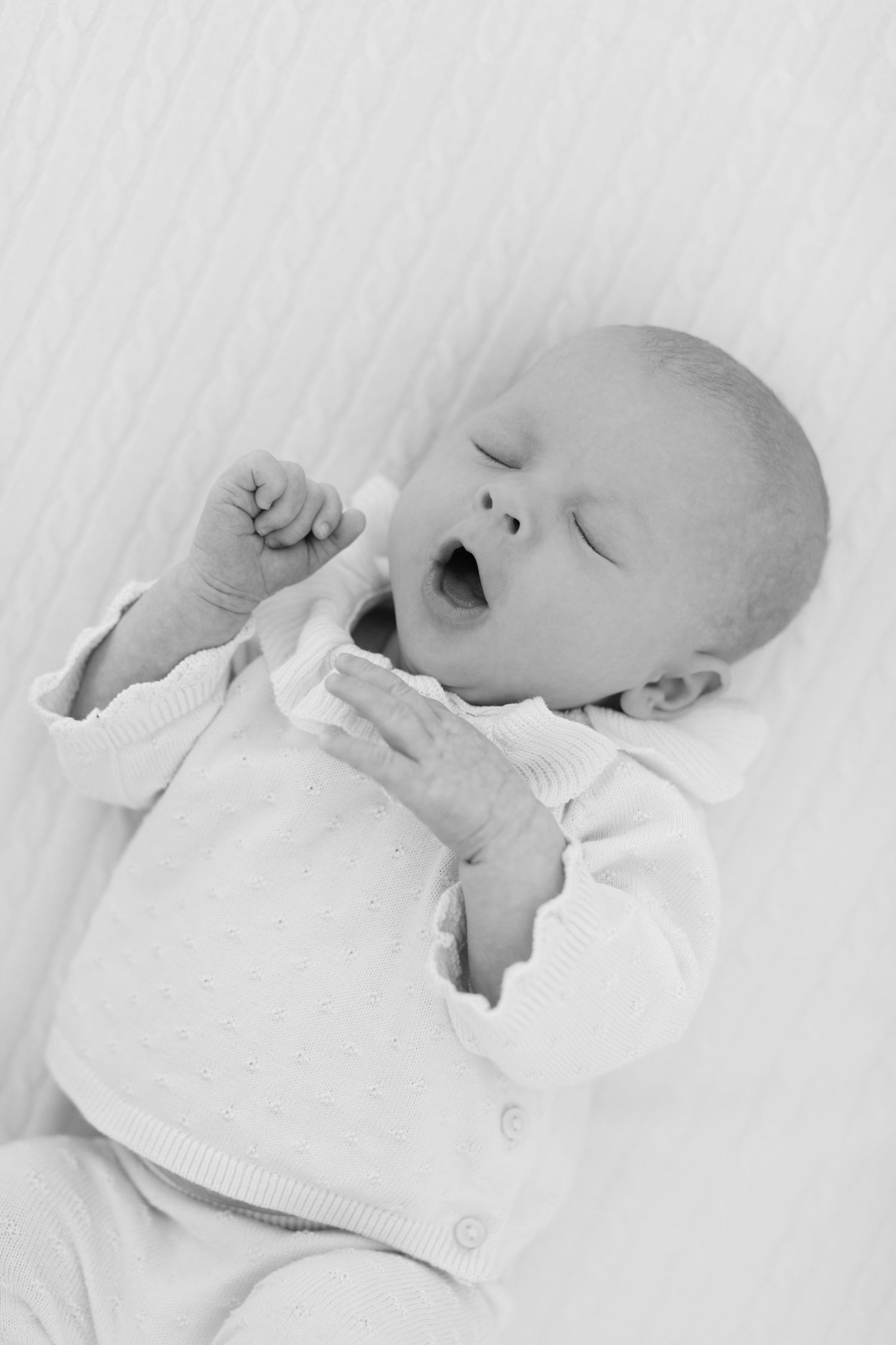 Black and white photo of a newborn baby yawning while lying on a white blanket wearing a soft white onesie.