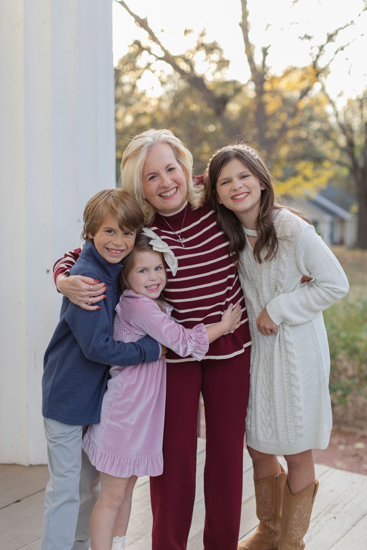 Three children share a joyful embrace while wearing casual fall clothing on a porch with autumn foliage in background.
