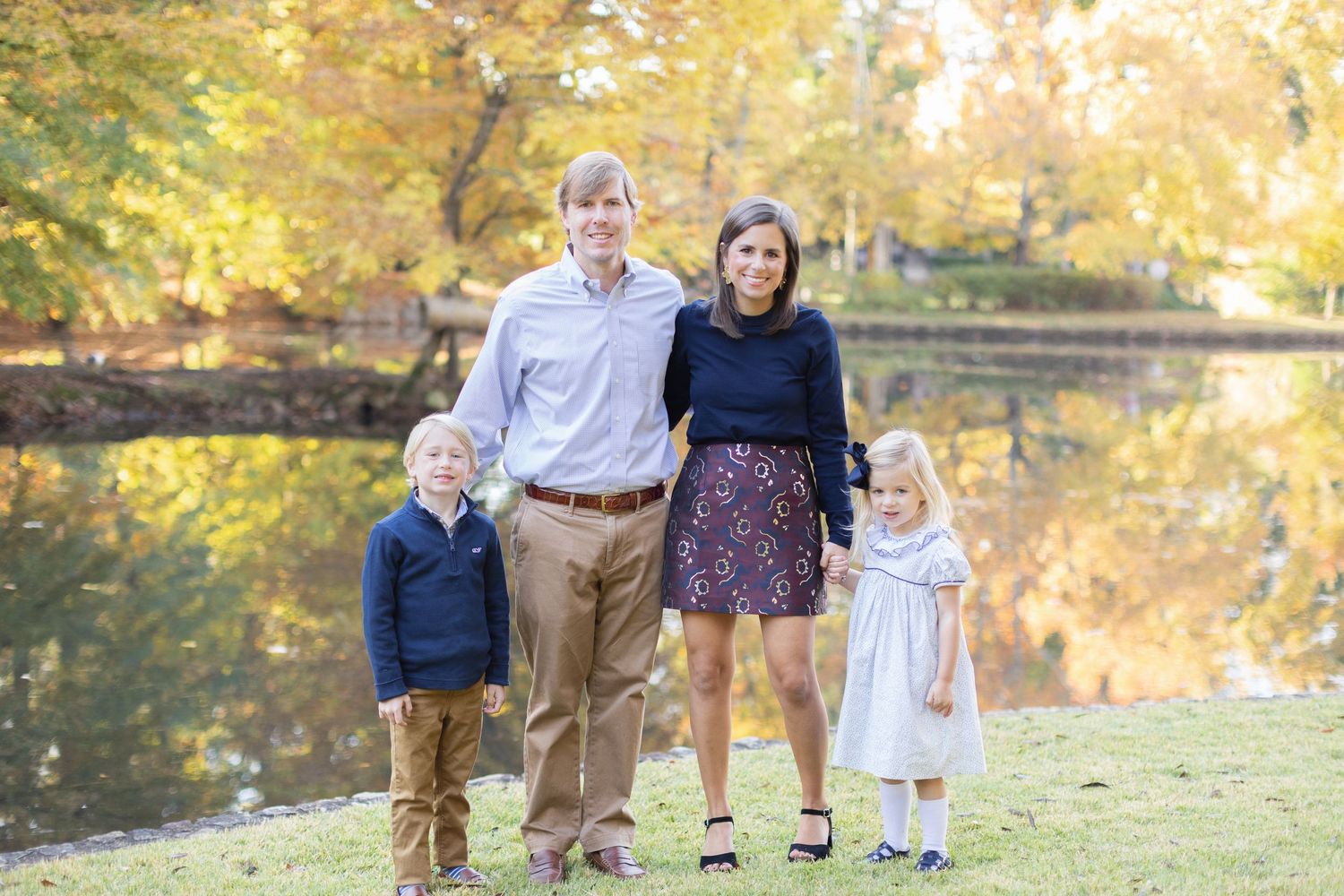 Family portrait session in a park during autumn with golden foliage and a pond in the background.