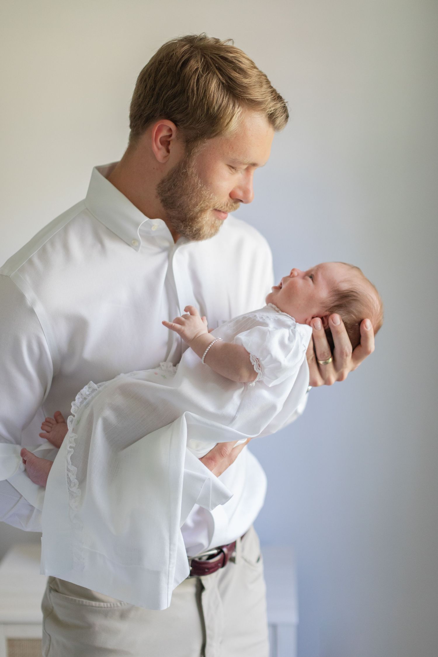 A bearded father in a white shirt tenderly holds his newborn baby dressed in white, gazing down lovingly.