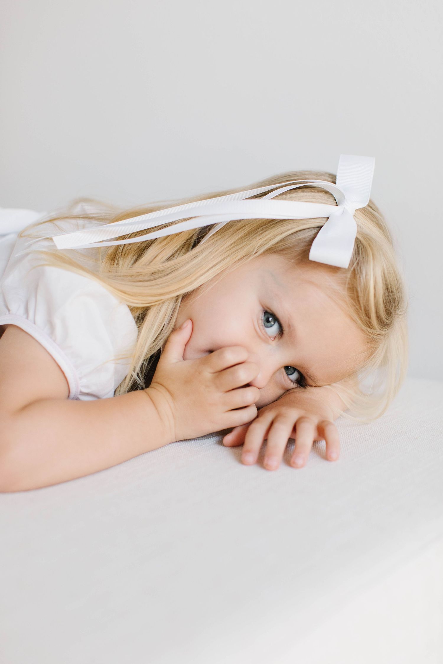 Child with blonde hair and white bow rests head on white surface in artistic portrait.