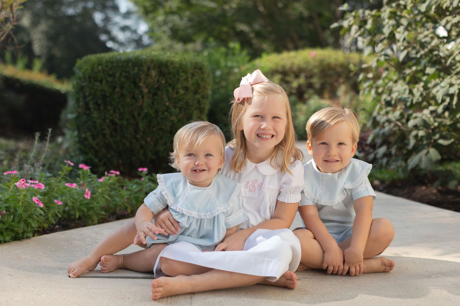 Three young children in light colored clothing sit together on a stone patio with a garden in the background.