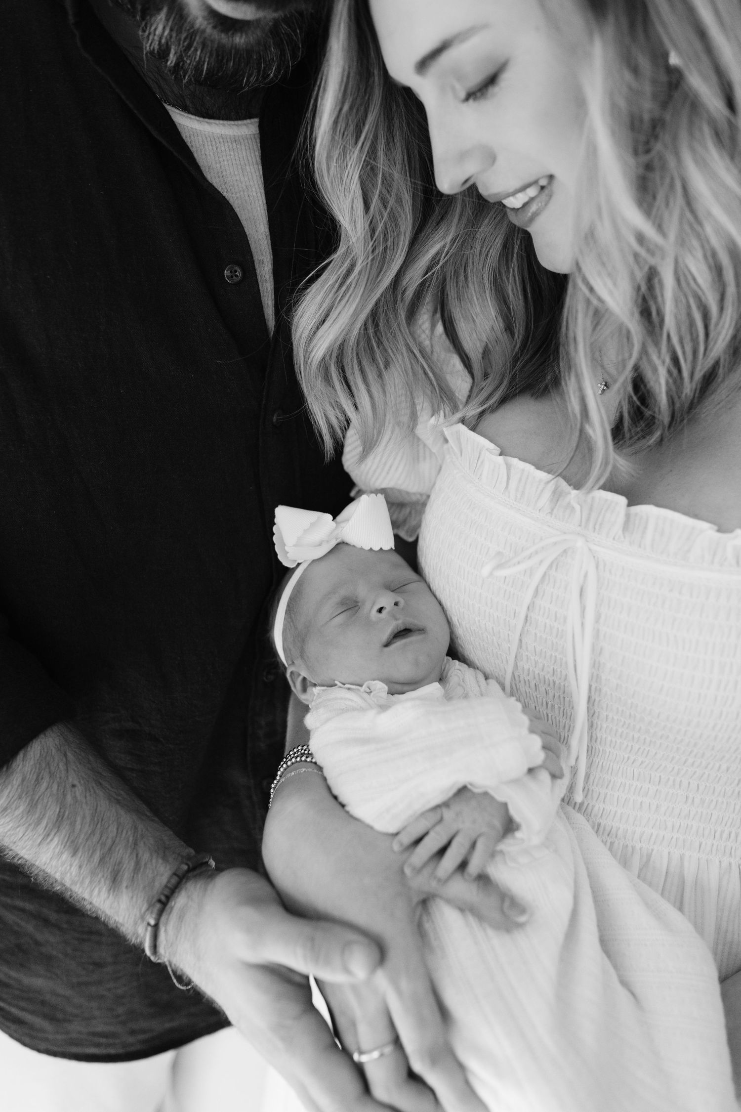 A black and white portrait of parents gazing lovingly at their newborn baby wearing a bow.