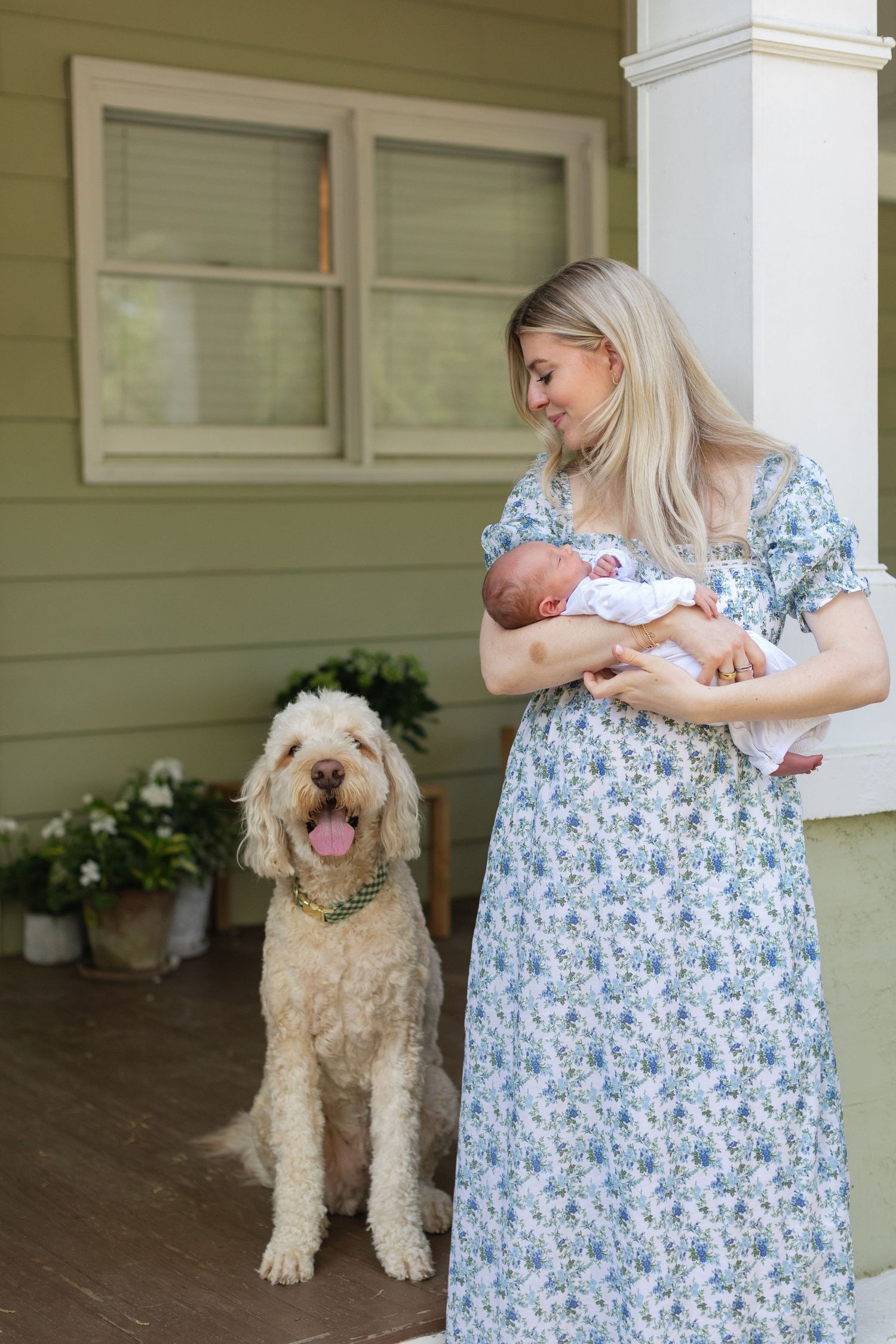 Blonde mother in floral dress holding newborn baby beside fluffy golden doodle dog on porch.