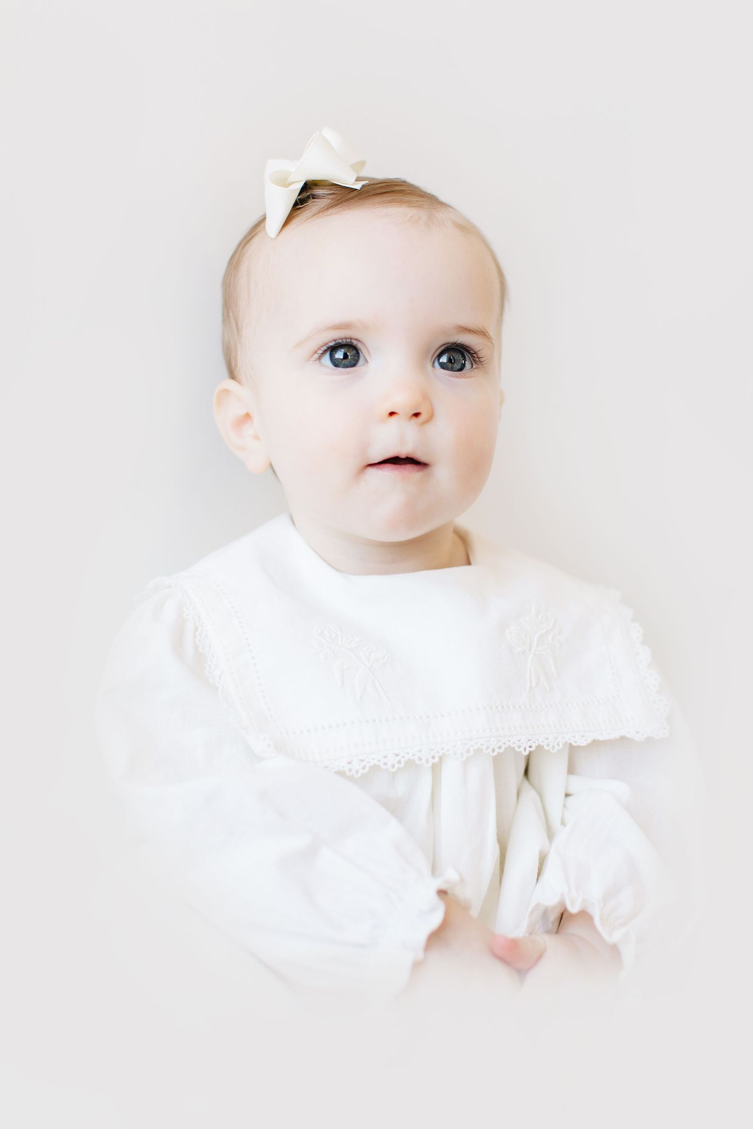 Adorable baby girl in white lace dress and bow headband with blue eyes looking up against white background.