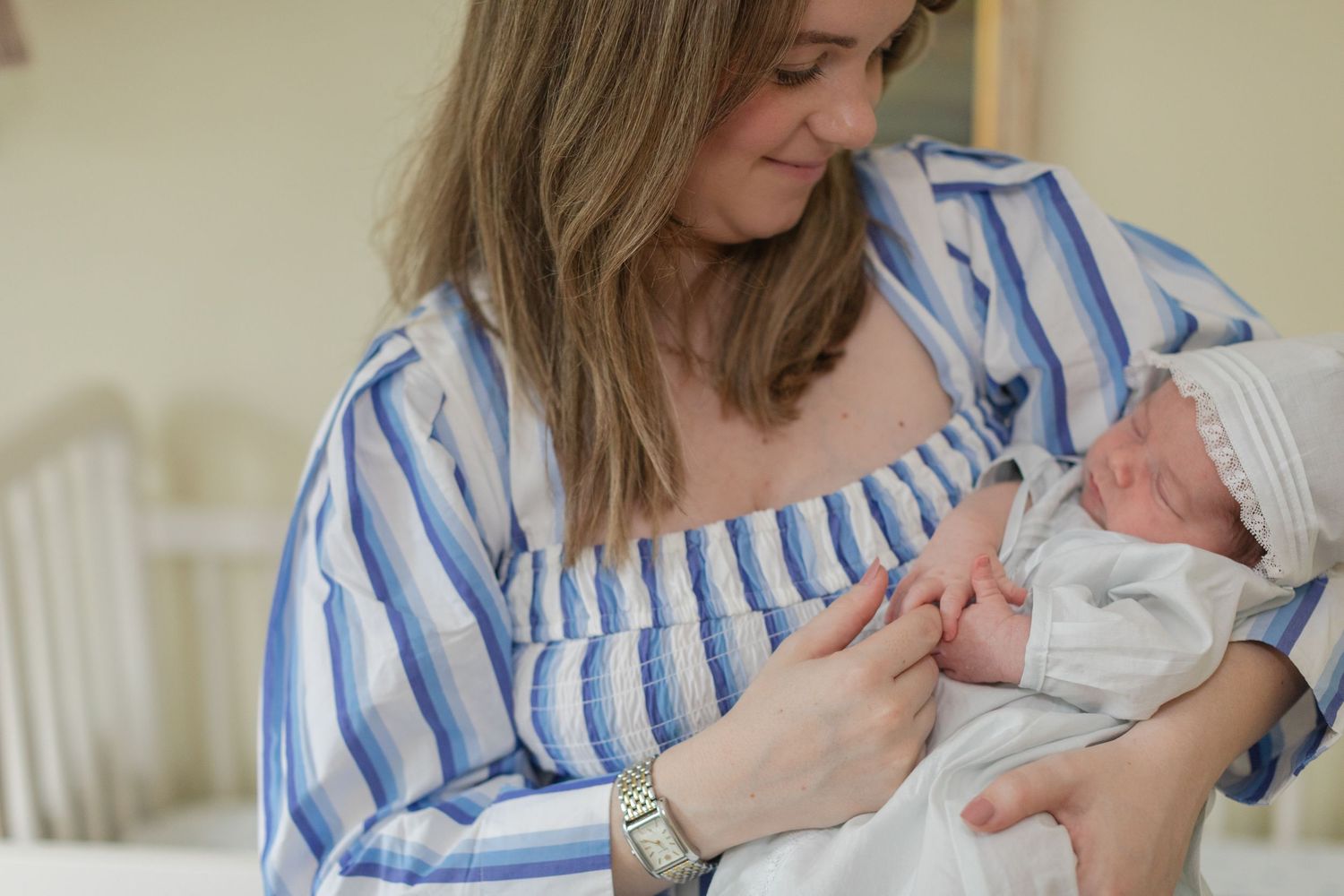 Tender moment captured as parent in blue stripes holds sleeping infant near nursery window.