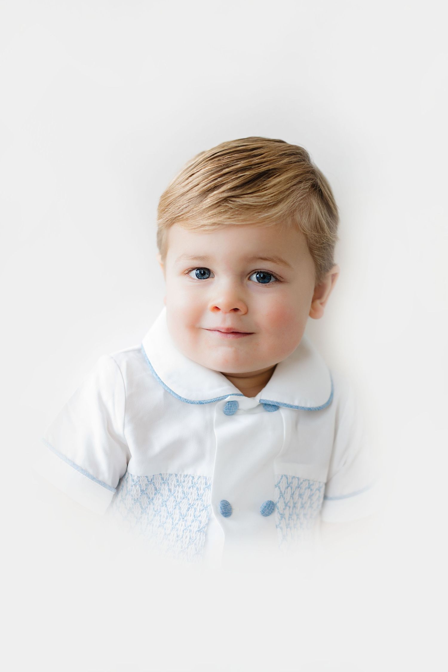 Sweet toddler wearing white and light blue collared shirt with smocked details smiles against white background.