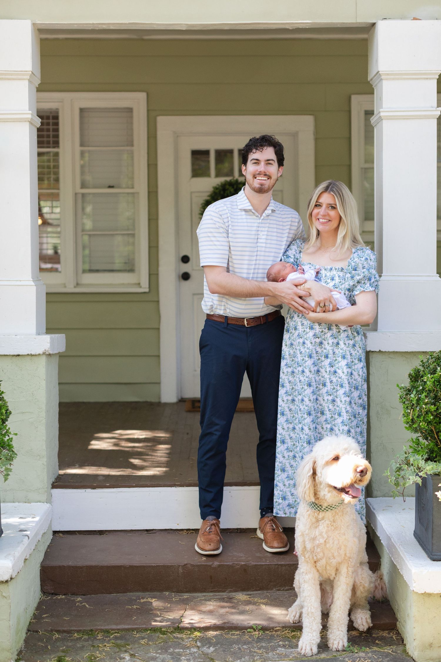 Young family with newborn baby and golden doodle dog posing on front porch of green home.