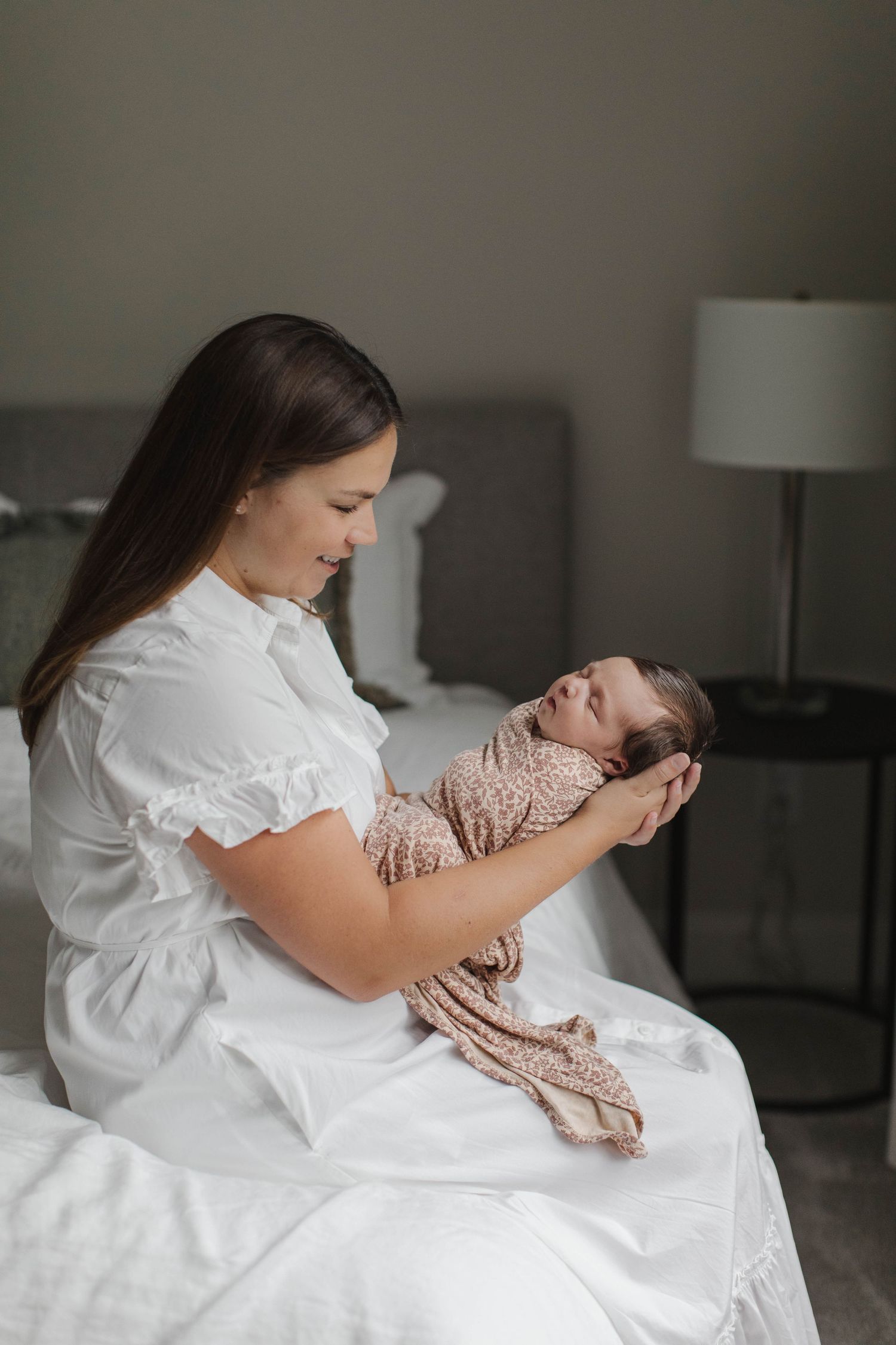 Mother in white dress holding sleeping newborn wrapped in blush swaddle in nursery.