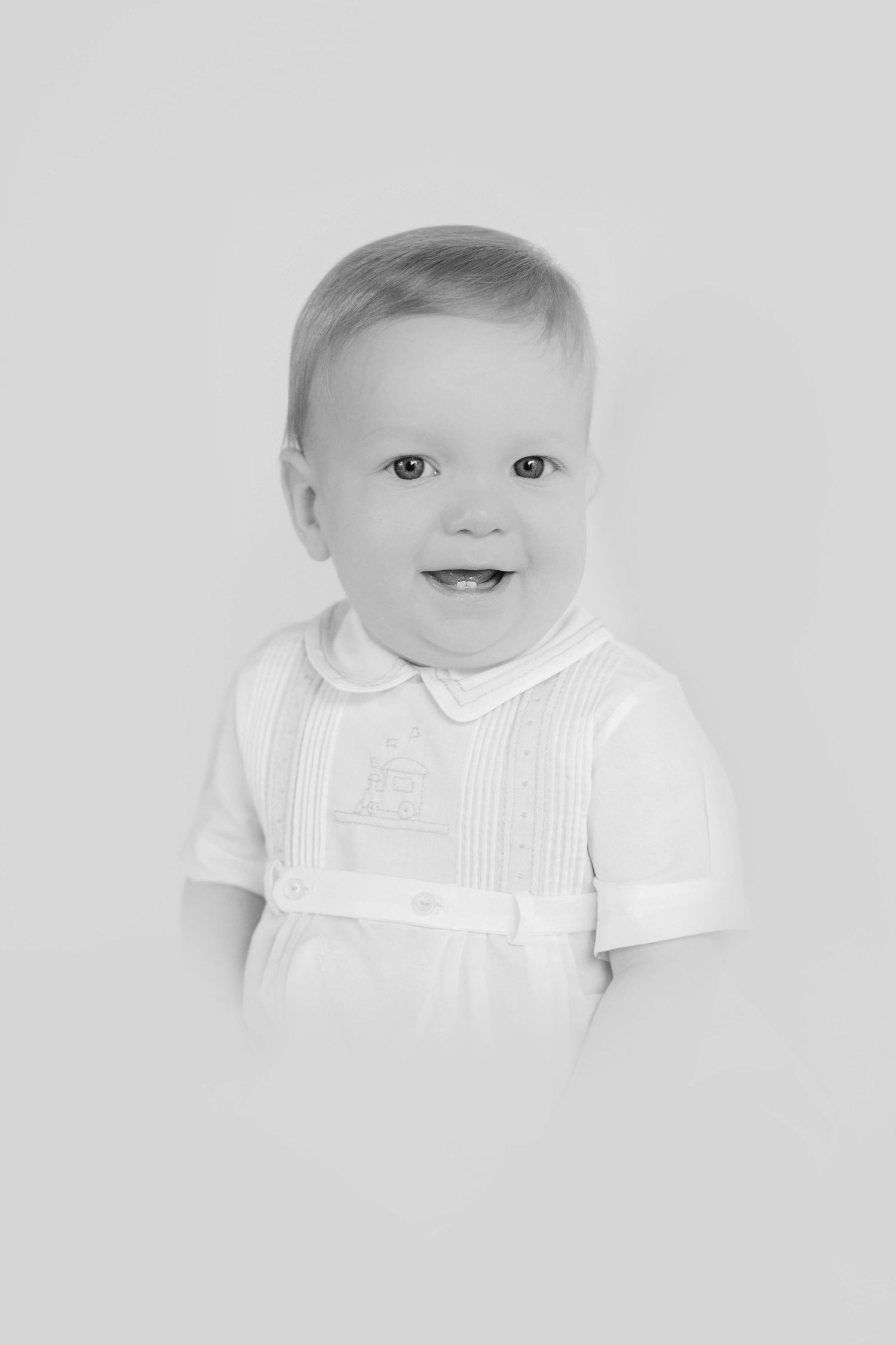 Black and white portrait of a baby in a white collared shirt with pleated detail and ribbon belt, smiling at camera.
