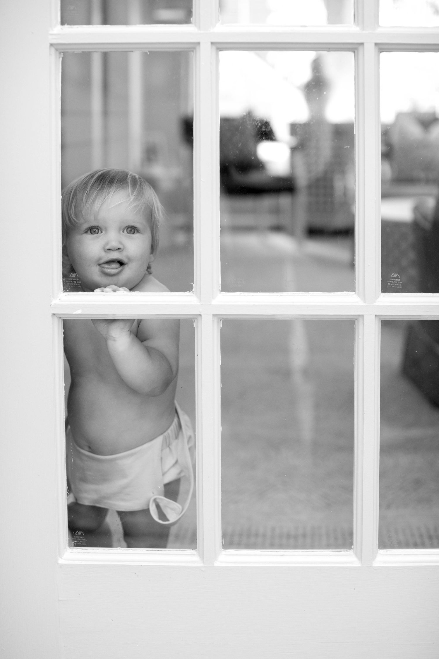 A small child peeks through white windowpanes with a joyful expression in black and white photography.