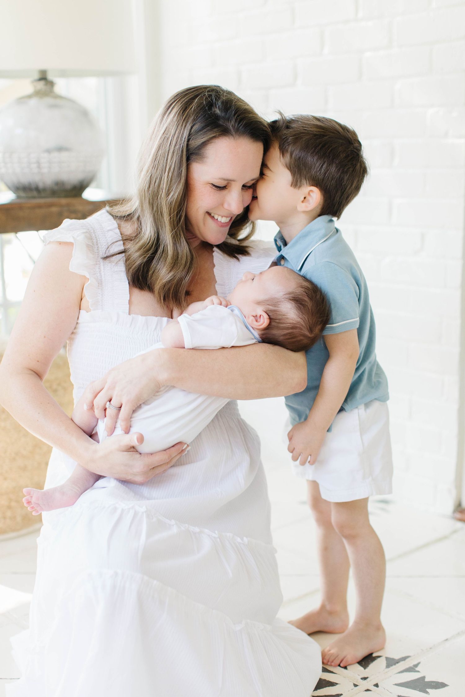 A tender family moment between mother, toddler and newborn in a bright white living room.
