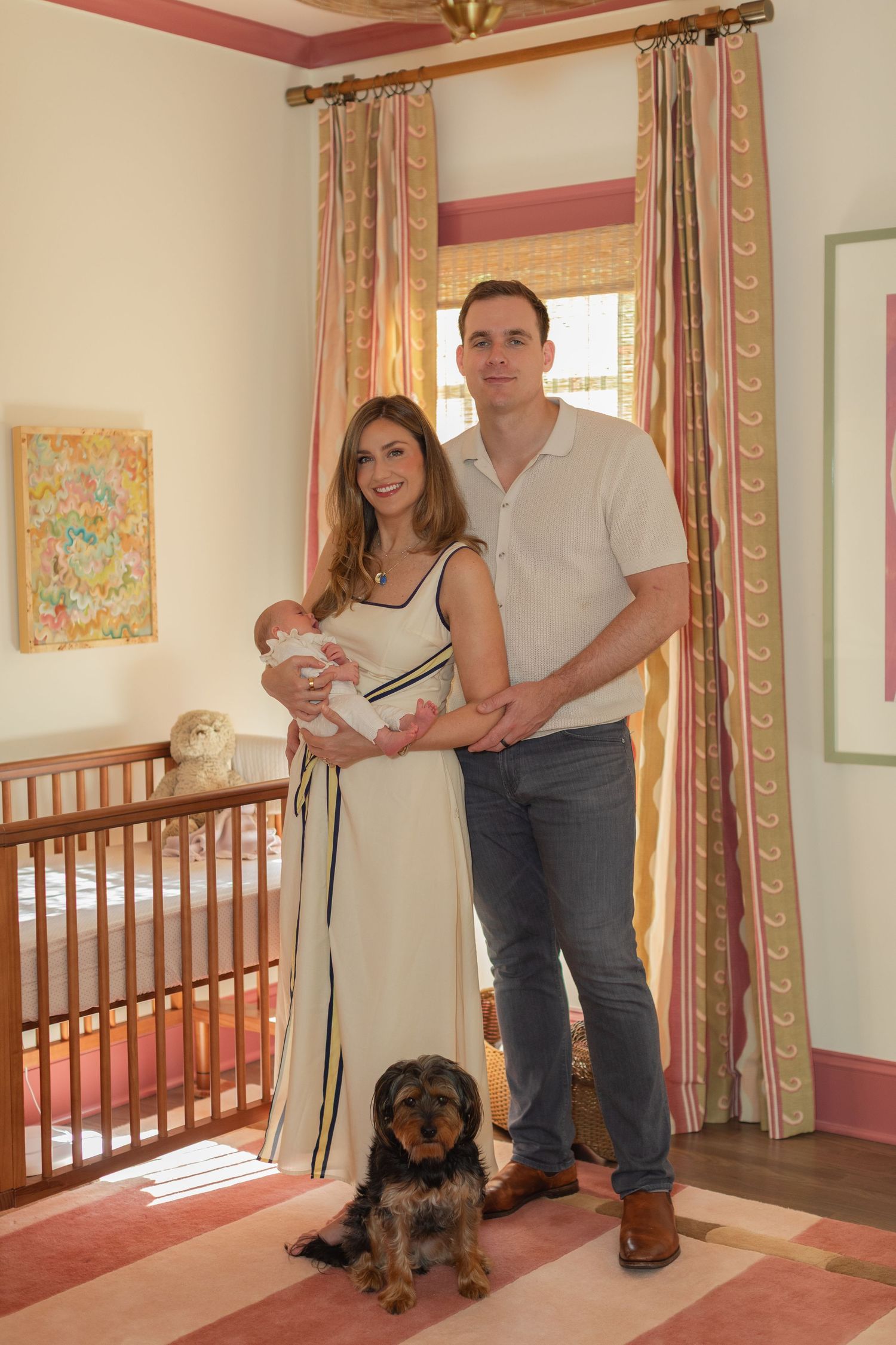 Young couple holding newborn baby with dog in decorated nursery room featuring pink curtains and wooden crib.