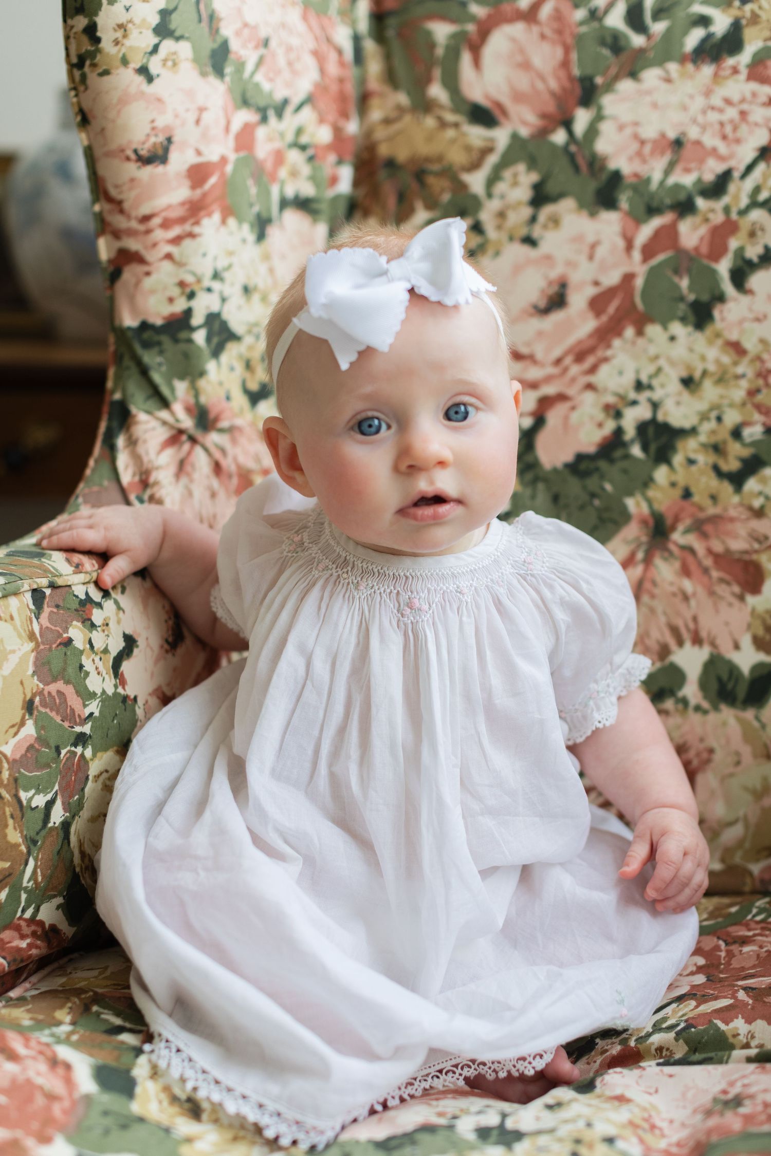 Baby girl in white smocked dress and bow headband sitting in floral armchair.