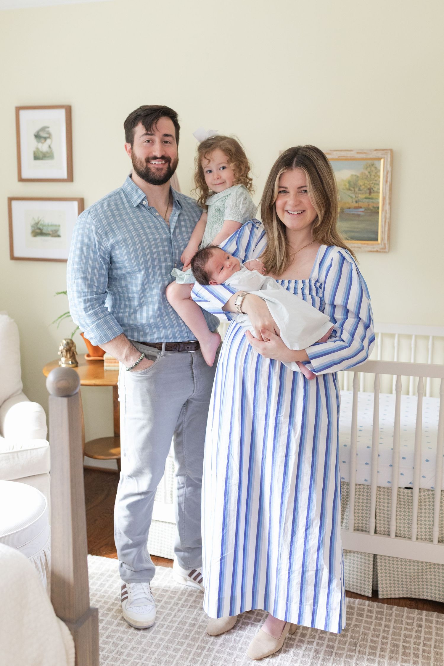 Family poses together in nursery room with newborn baby, wearing coordinating blue and white striped outfits.