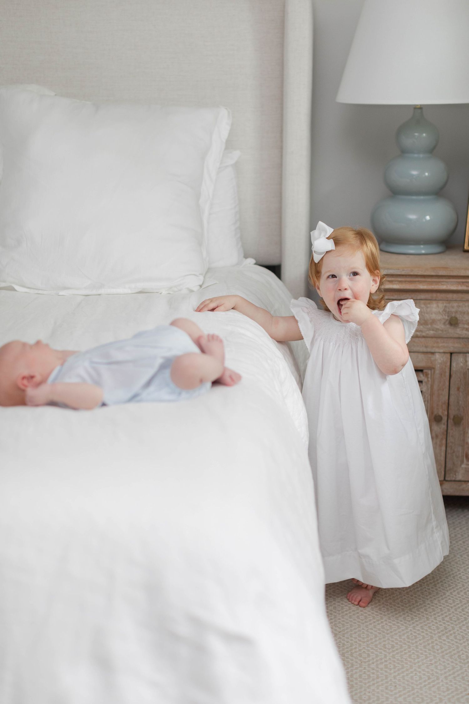Toddler in white dress stands beside bed with sleeping baby while light blue ceramic lamp adorns bedside table.