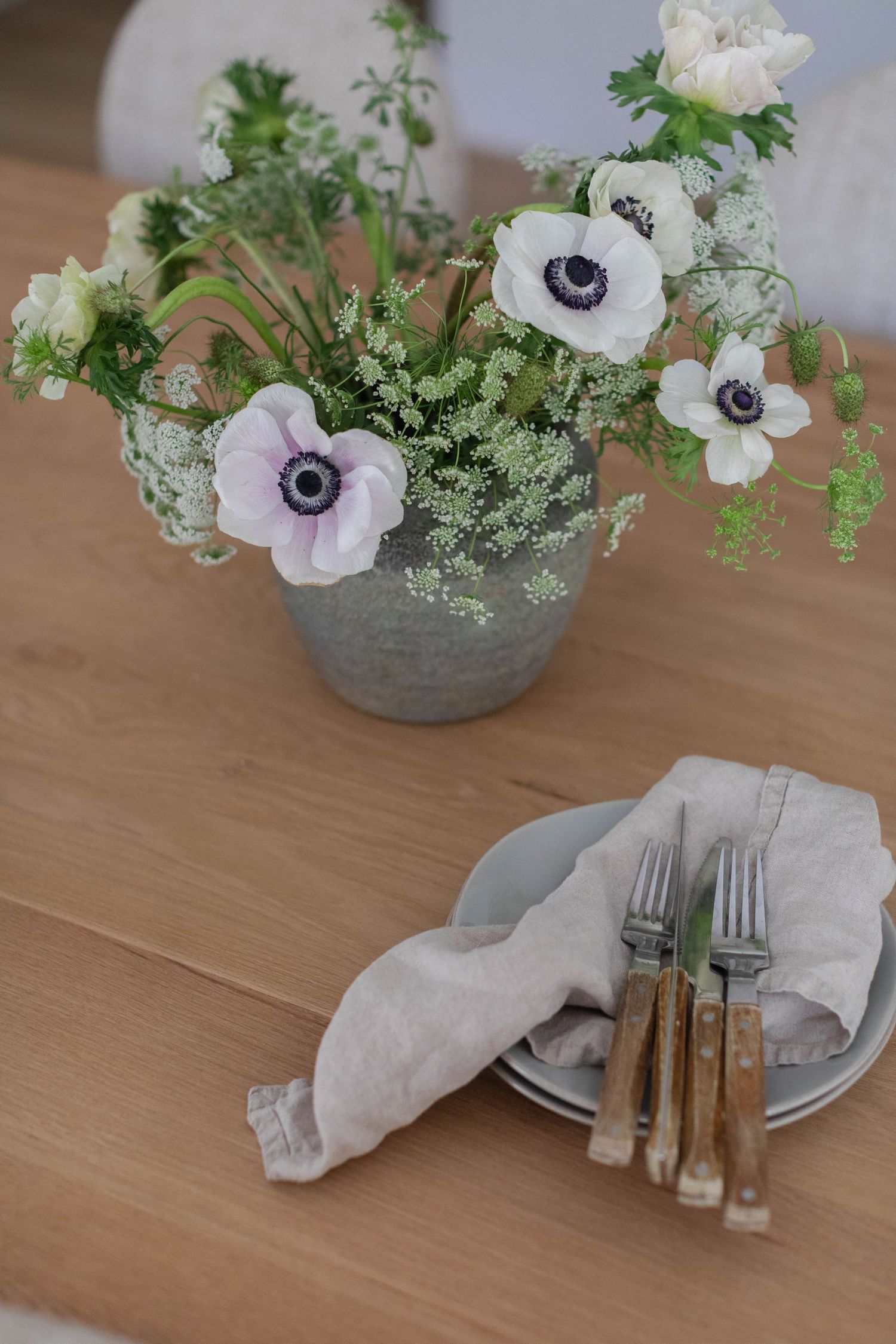 White anemone flowers in a gray pot with ferns alongside rustic wooden-handled cutlery and linen napkins on wooden table.