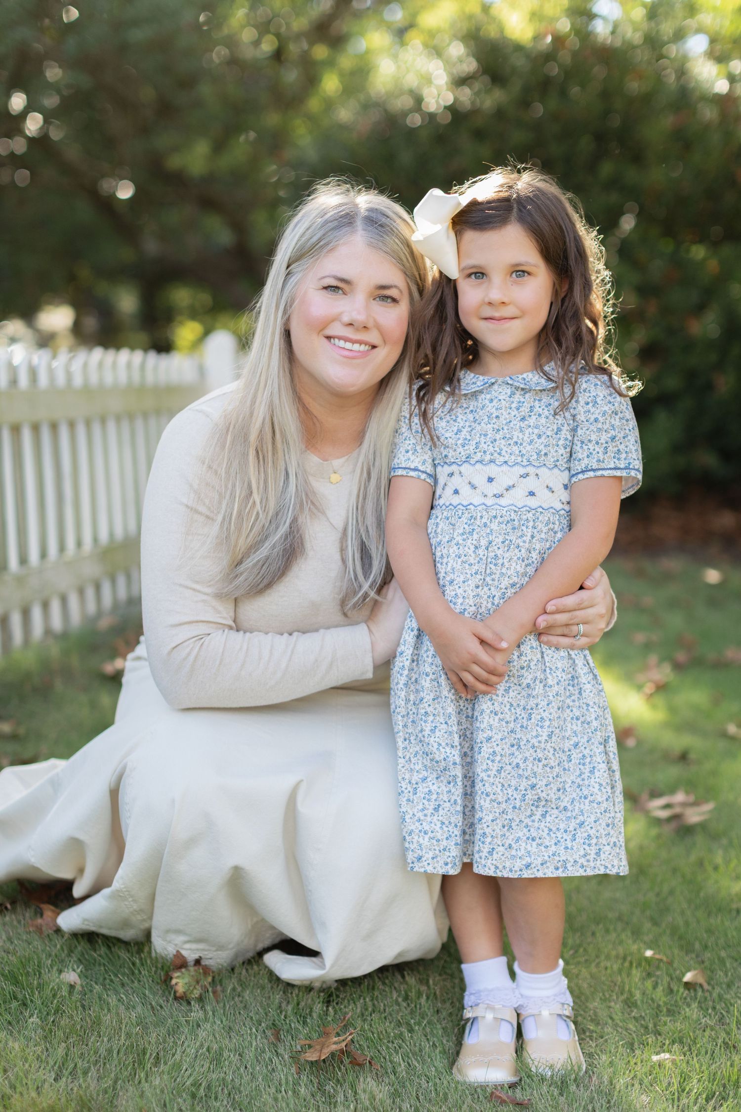A sweet outdoor photoshoot showing a parent and child in coordinating cream and floral dresses sitting on grass.