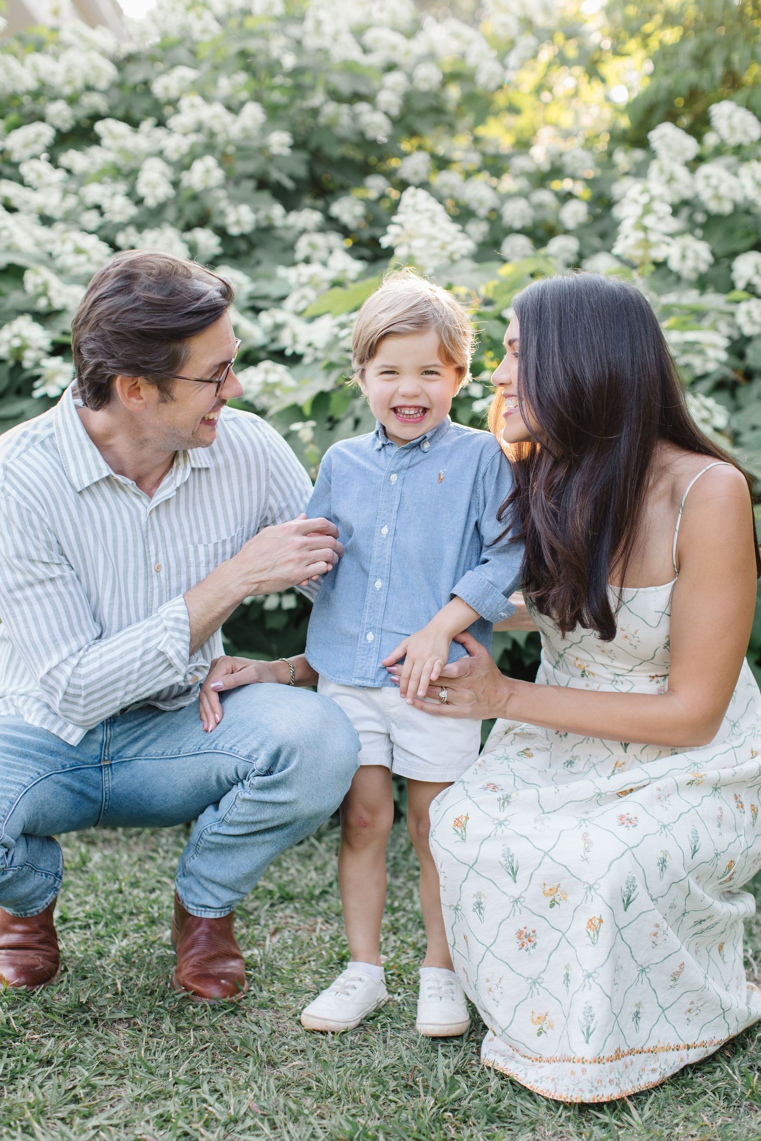 A happy family with a laughing toddler boy poses outdoors near white blooming bushes in a bright garden setting.