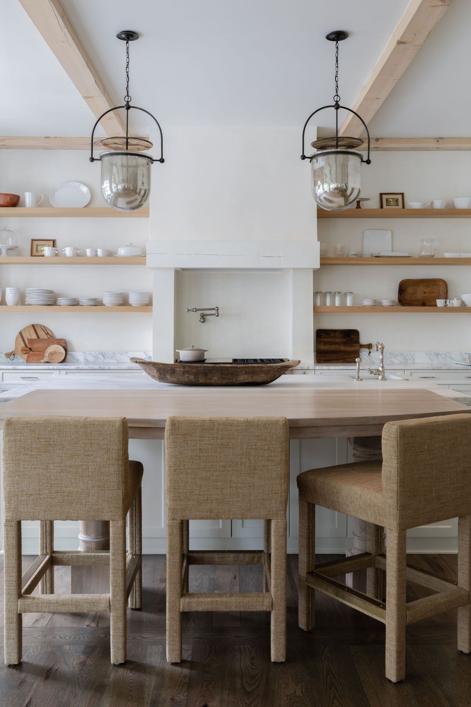 Bright farmhouse kitchen with wood island, linen barstools, open shelving, and two glass pendant lights over white walls.