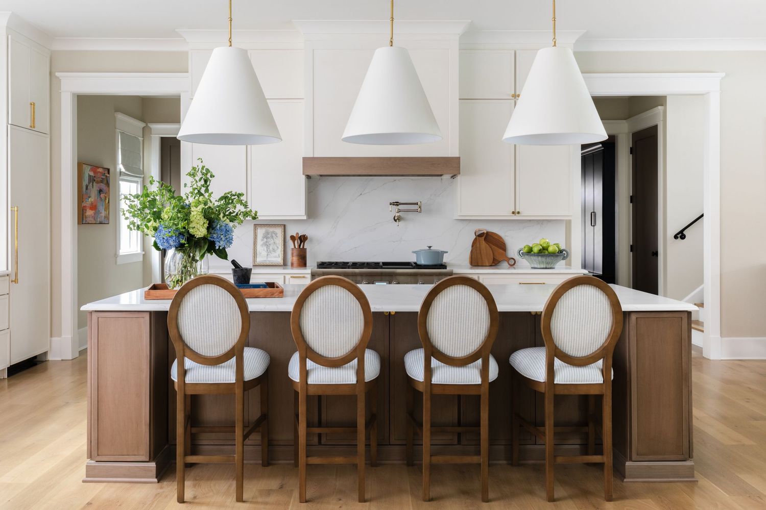 Modern white kitchen with wood island, upholstered round-back bar stools and pendant lighting over herringbone floors.